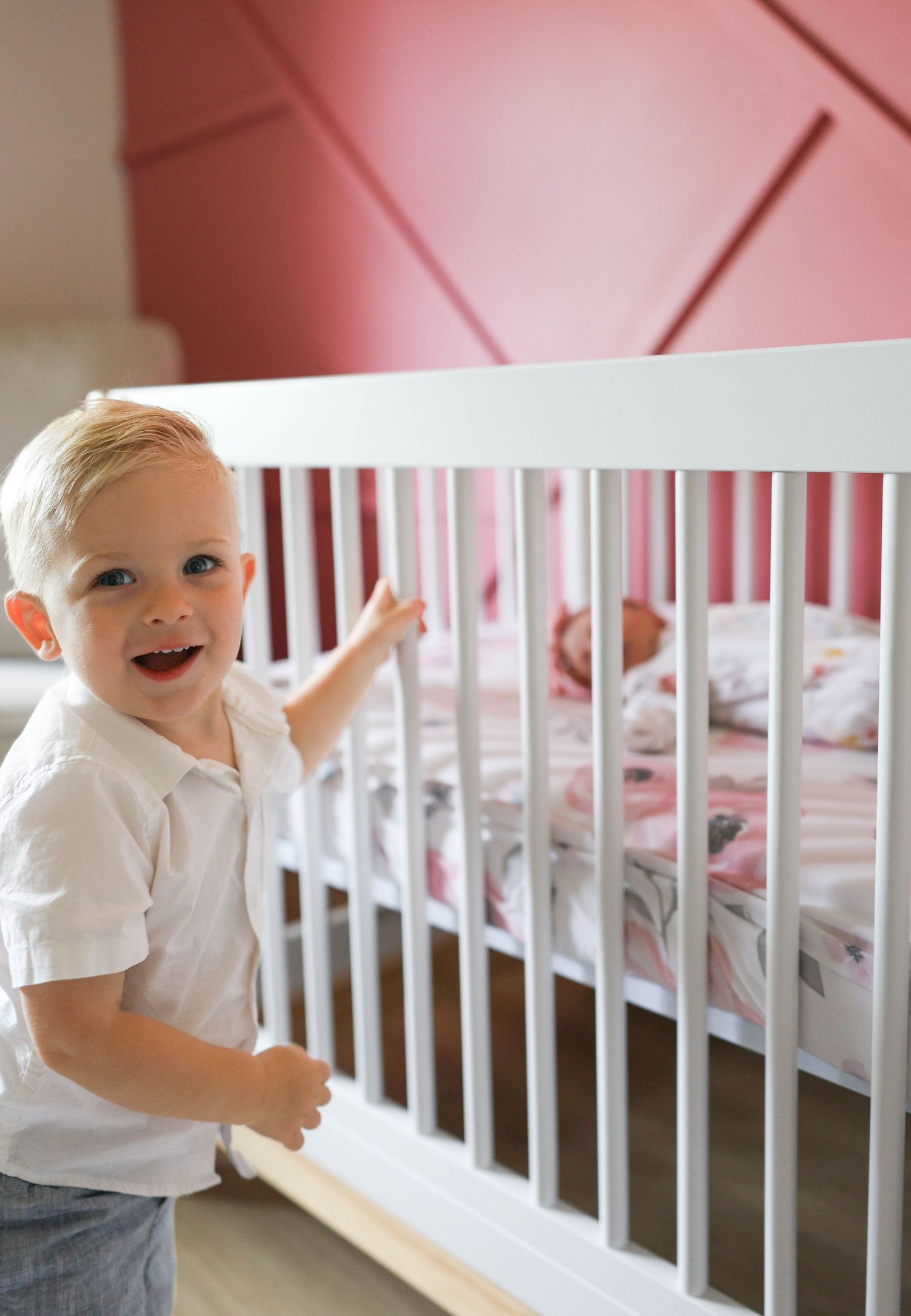 Smiling toddler boy in white shirt standing beside a white crib in a pink nursery room.