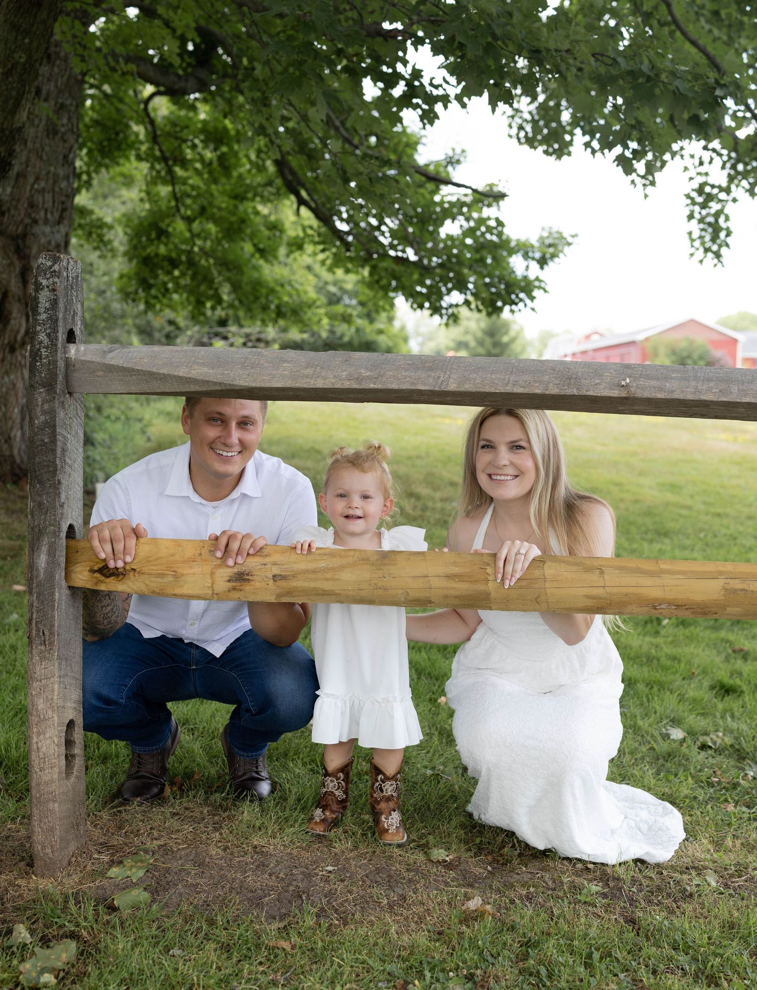 Happy family with toddler posing behind rustic wooden fence rail in lush green park setting.