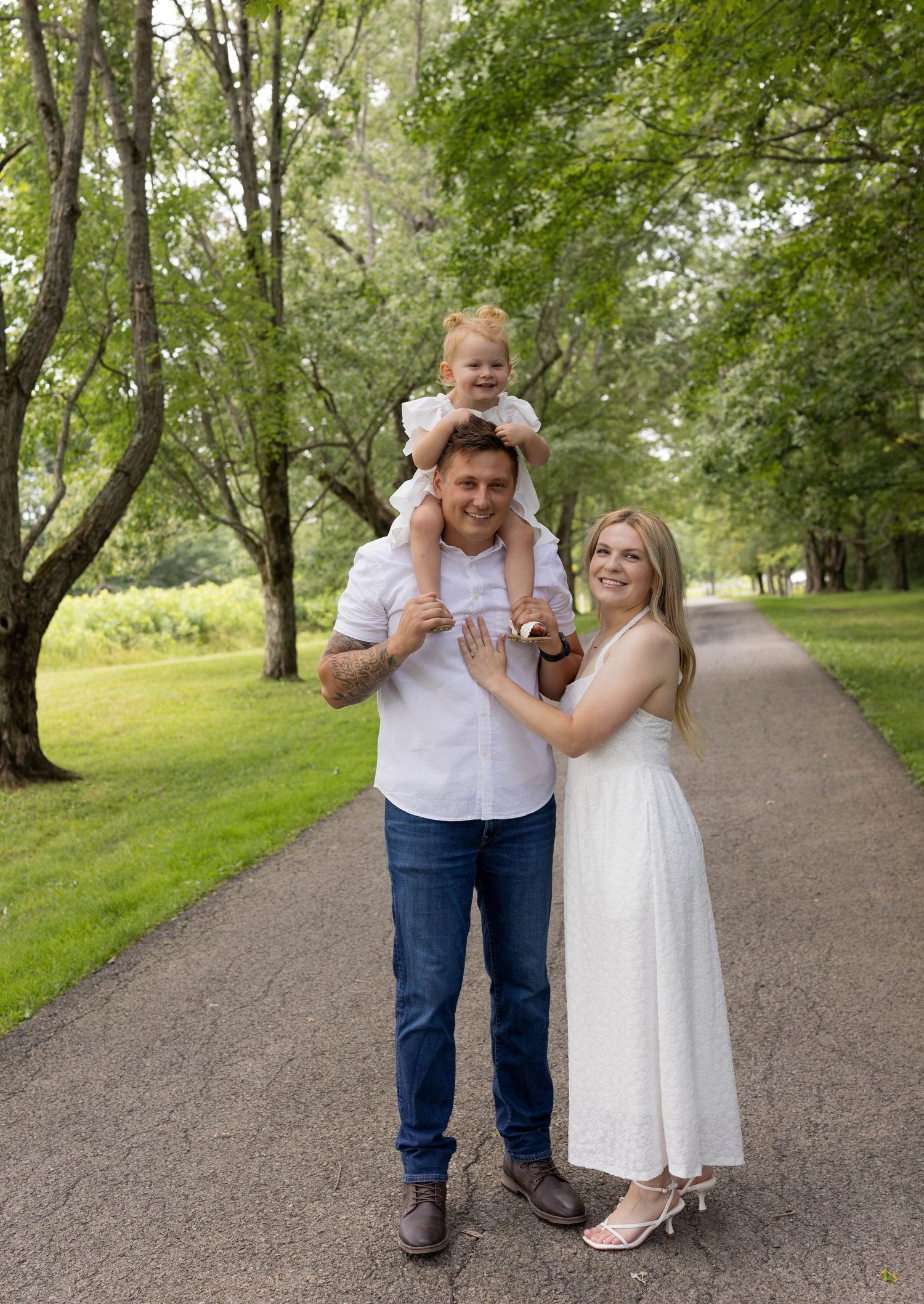 Happy family of three poses on a scenic tree-lined path, with toddler sitting on dad's shoulders.