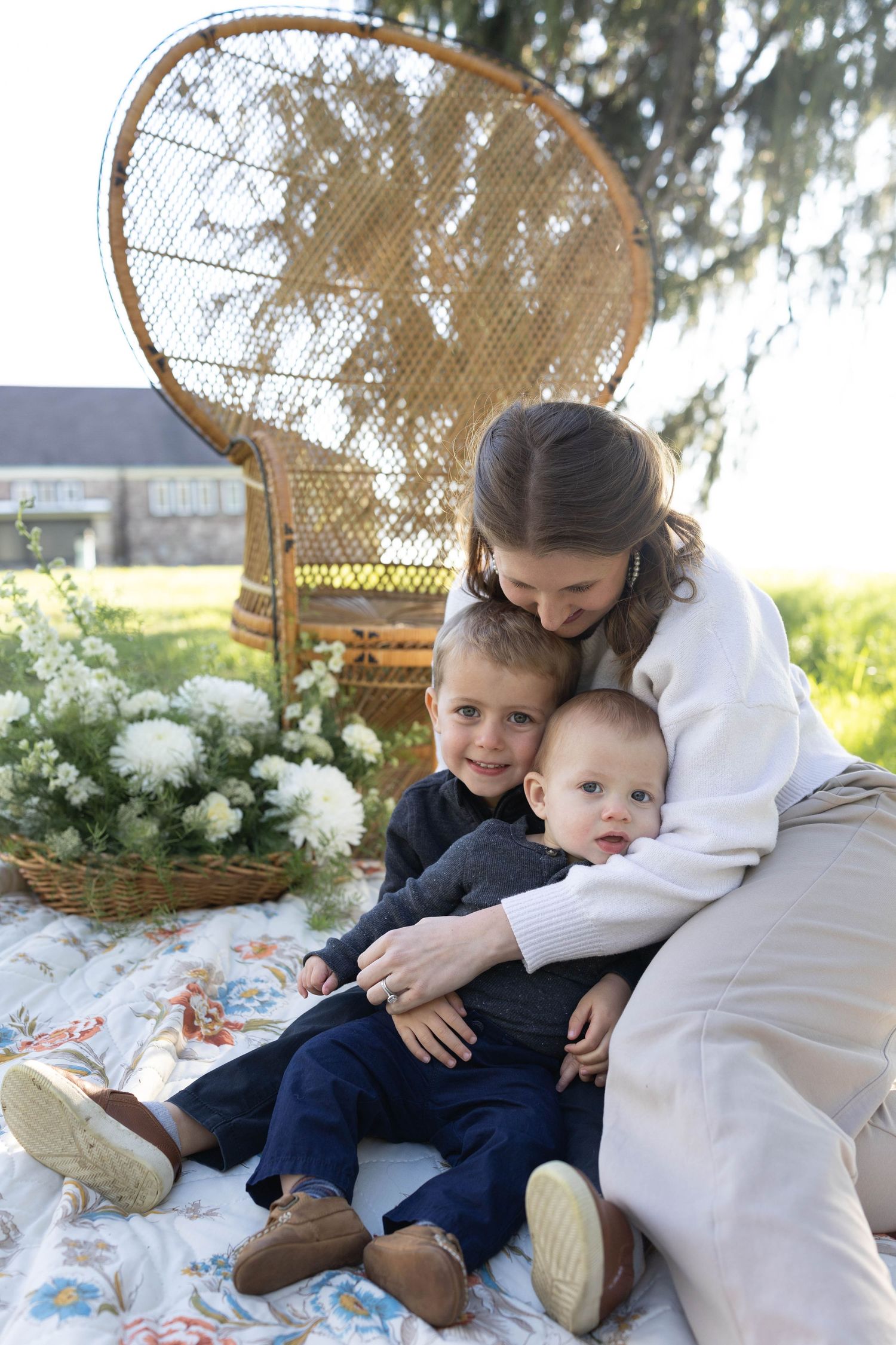 Peaceful outdoor scene with wicker peacock chair and family sitting on floral blanket in garden with white flowers.