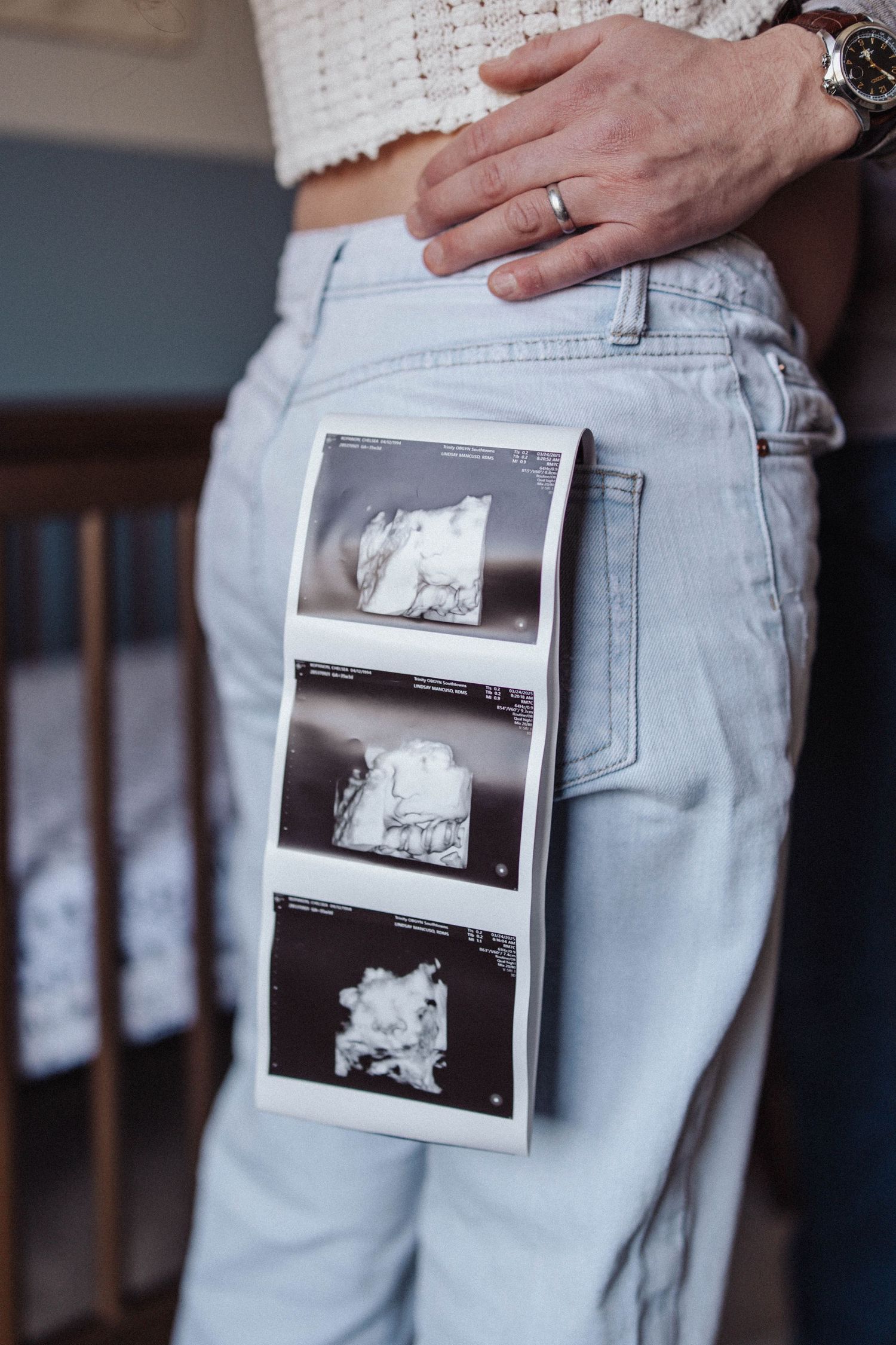 A strip of ultrasound photos tucked into the pocket of light wash denim jeans with a hand wearing a wedding ring visible.