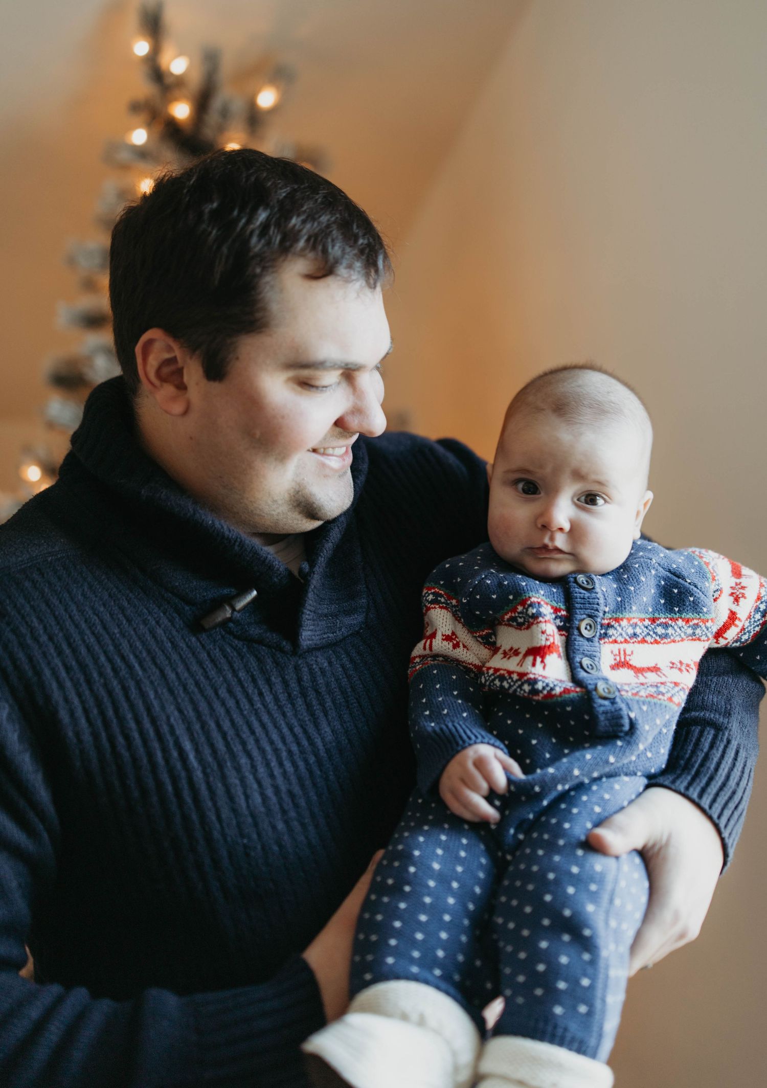 Adorable baby in polka dot outfit and winter sweater being held while Christmas lights twinkle in background.