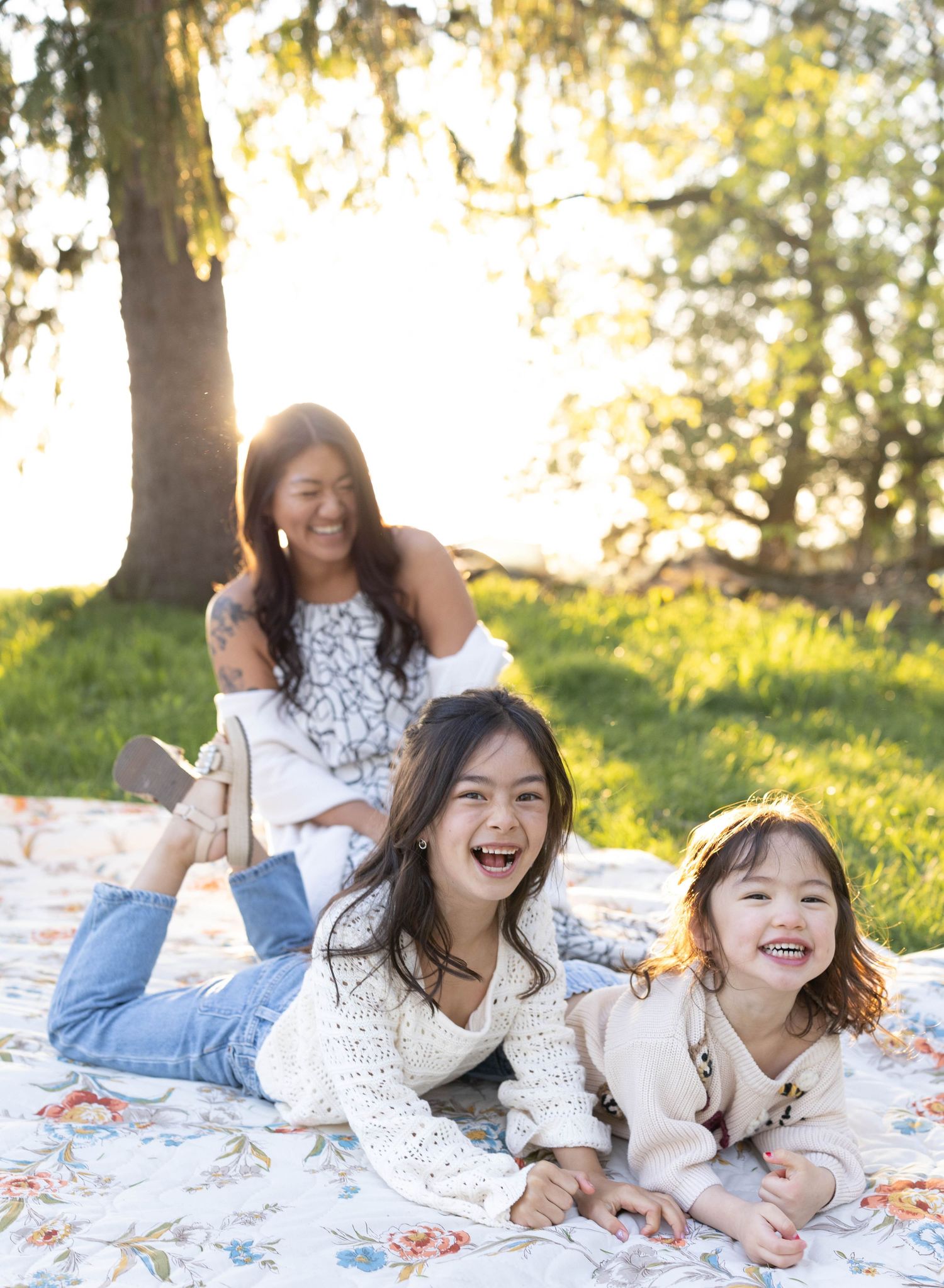 Three people lying on a floral blanket outdoors on grass enjoying a sunny afternoon together.