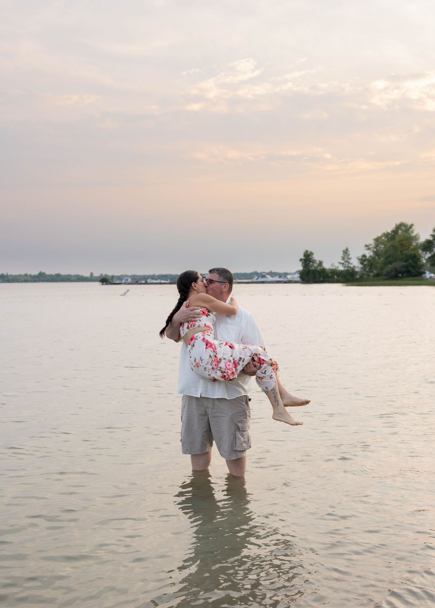 A couple shares a romantic kiss as the man carries the woman in shallow lake water at sunset.