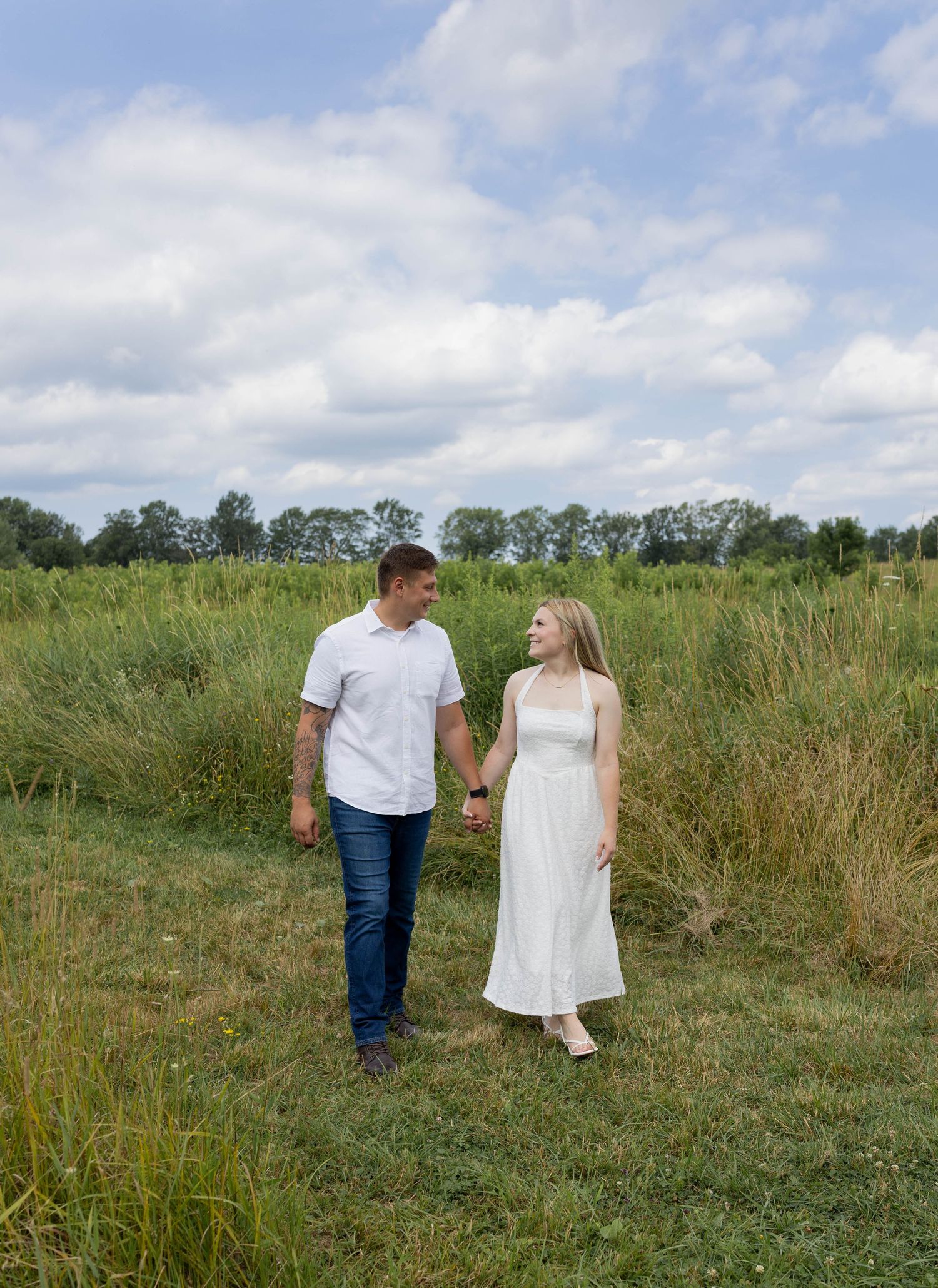 Couple holding hands walking through a lush green field under a cloudy sky, woman in white dress, man in jeans.