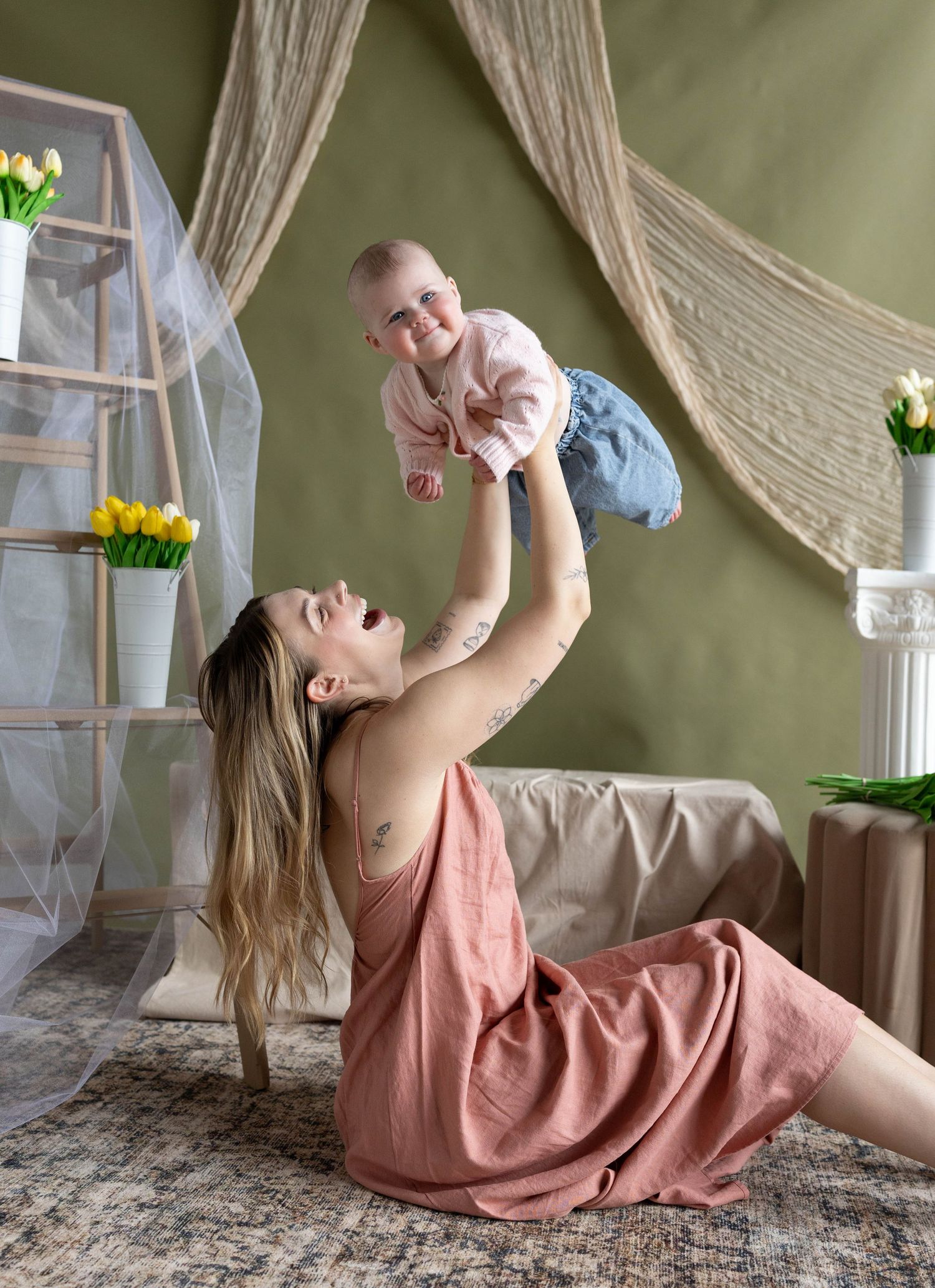 A joyful mother in a pink dress lifts her smiling baby overhead while sitting on a rug in a green-walled room.