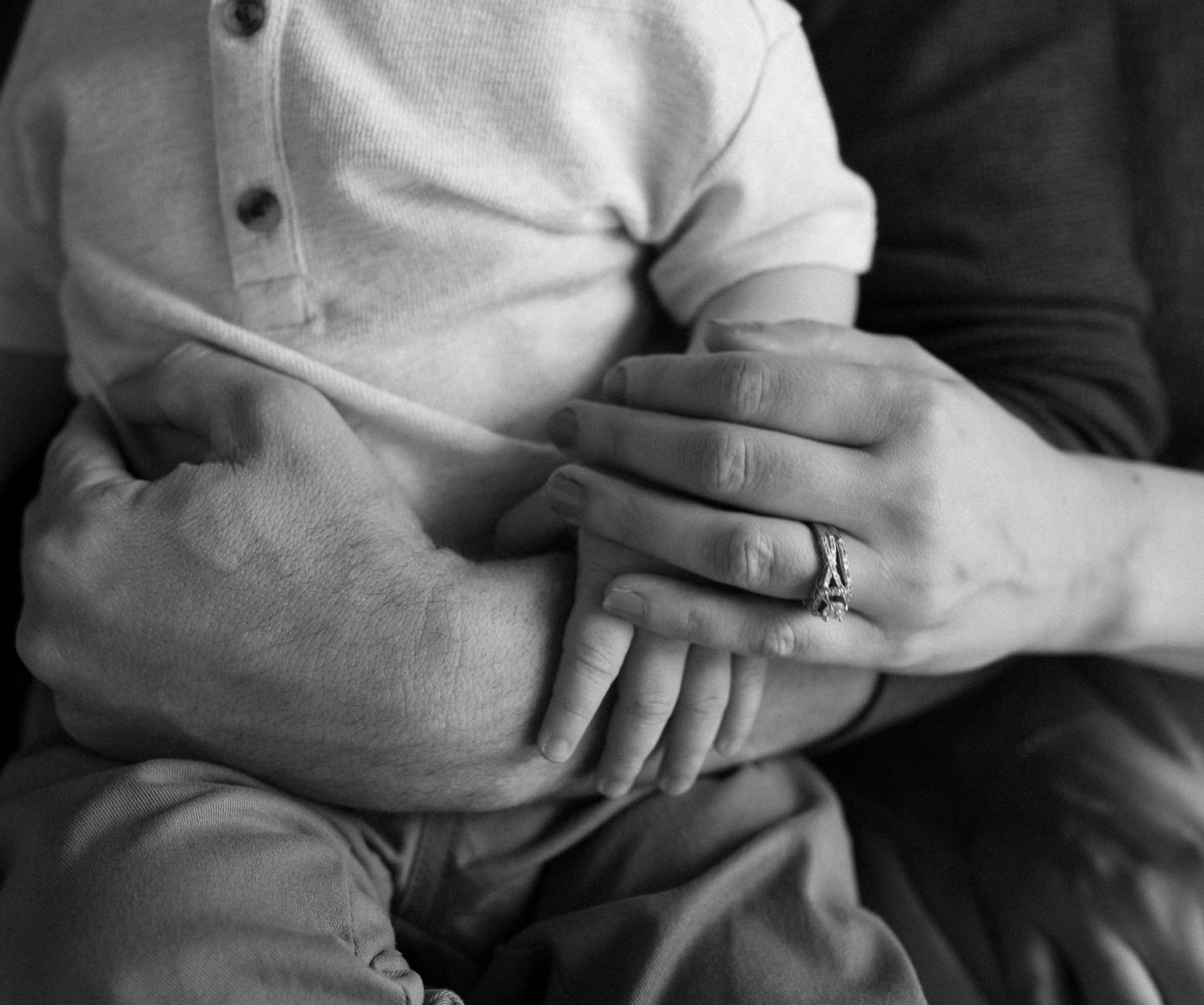 Black and white photo of a couple holding hands, woman wearing a ring, with a baby resting in their arms.