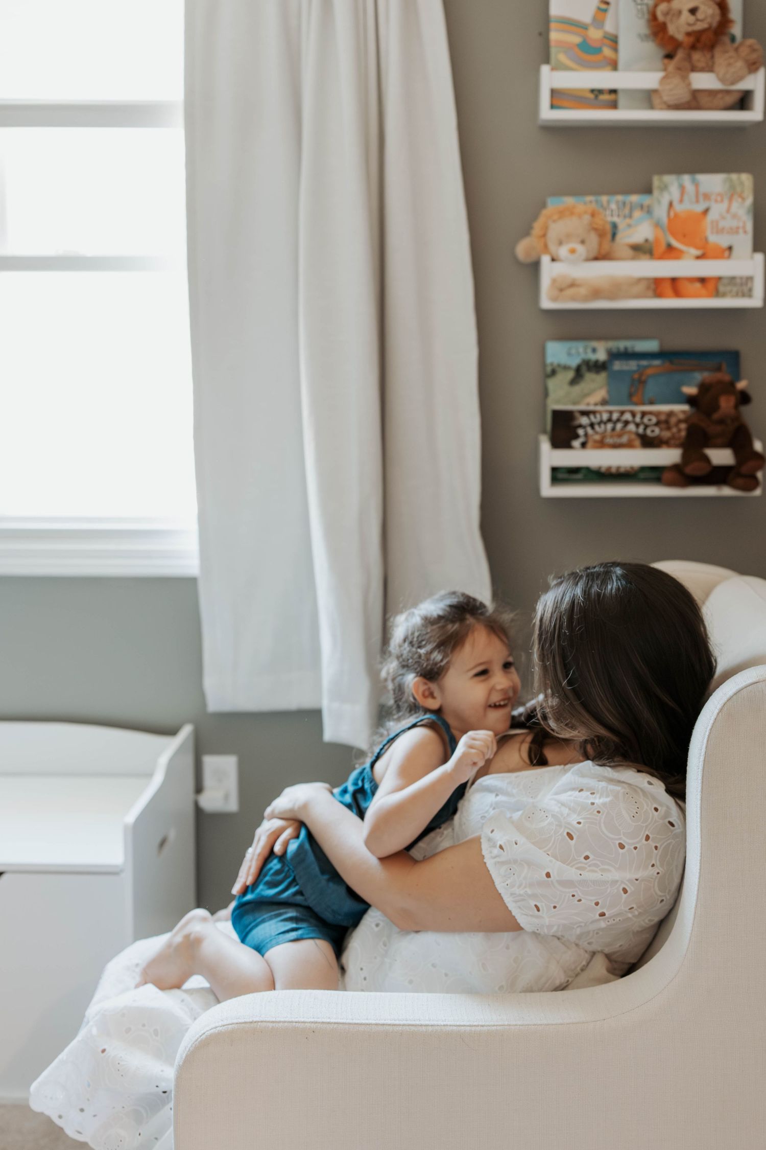 Mother sitting in nursery glider chair cuddling toddler girl, with children's books on wall shelves behind them.