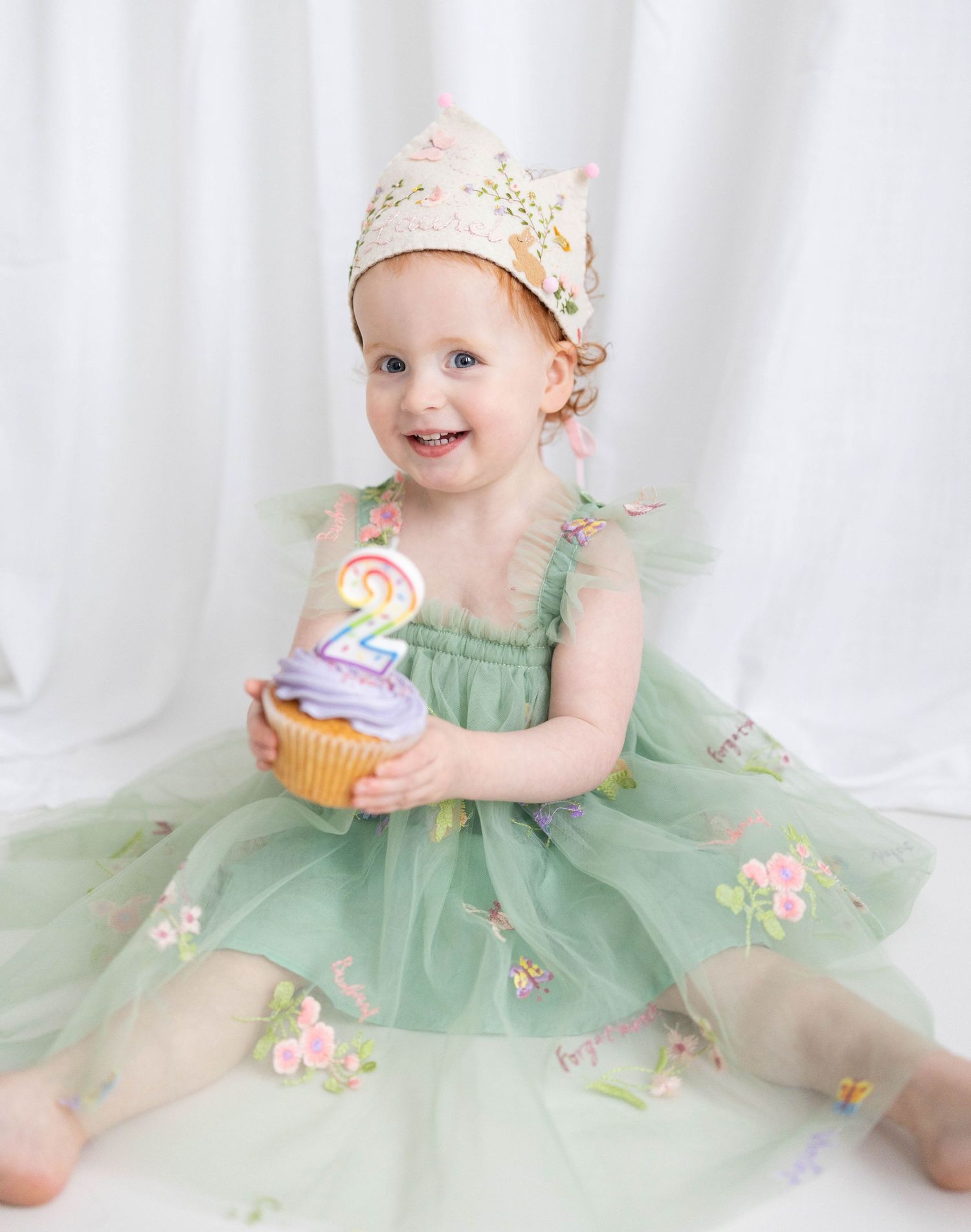 Sweet child in a mint green tulle dress and floral crown holds a birthday cupcake with number 2 candle.