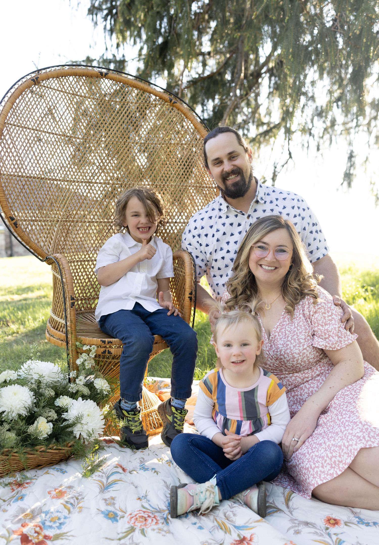 Family sits together on a floral blanket with decorative wicker peacock chair and white hydrangea flowers outdoors.