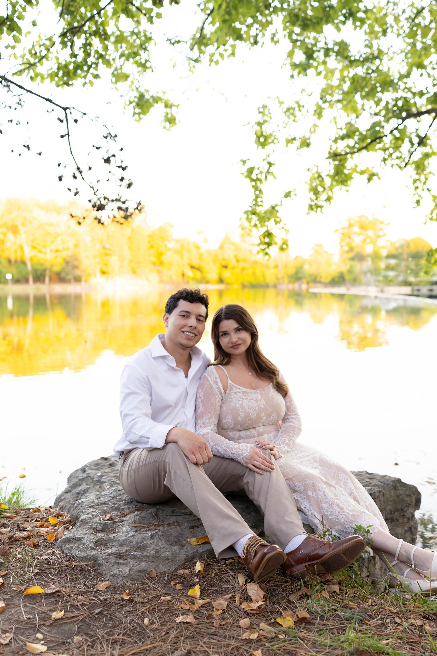 A couple sits together on a large rock by a tranquil lake with autumn foliage reflecting in the golden water.