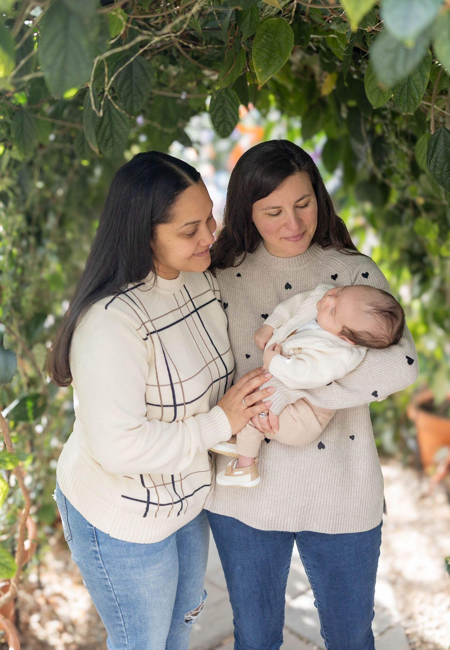 Two women in sweaters standing under green foliage admire a sleeping baby wrapped in white.