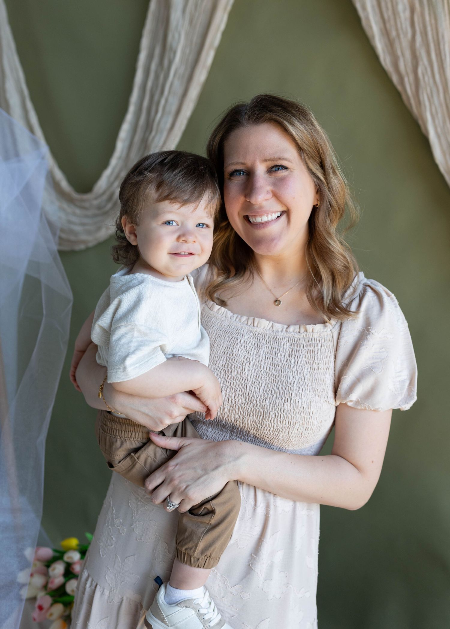 Smiling woman in beige dress holding a happy toddler boy, posing together in front of a green backdrop.