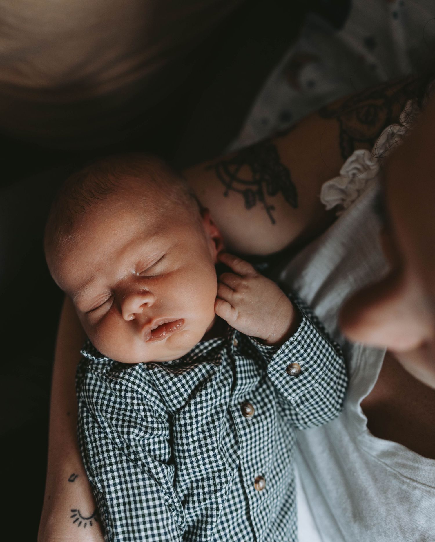 A sleeping newborn in a gingham shirt rests peacefully in a tattooed parent's arms.