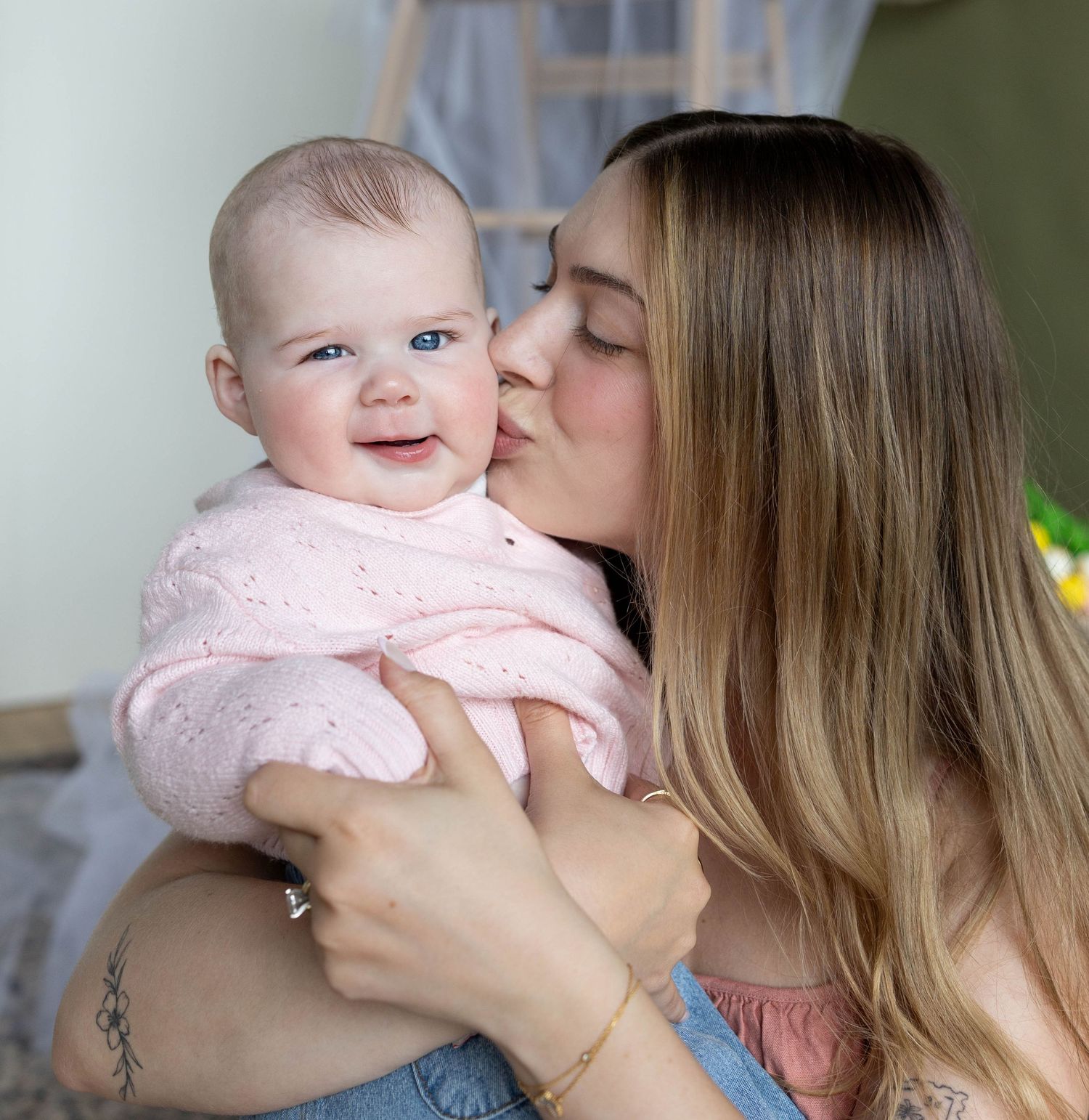 Mother kissing smiling baby girl in pink knit sweater while holding her in arms.