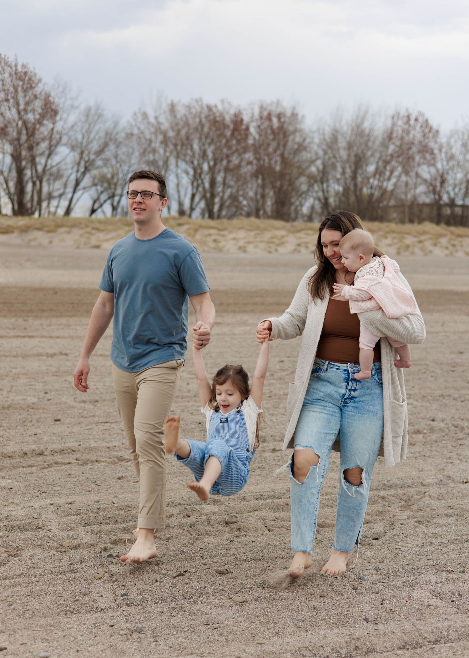 A young family walks barefoot on a sandy beach, with dad swinging a toddler girl while mom holds a baby.