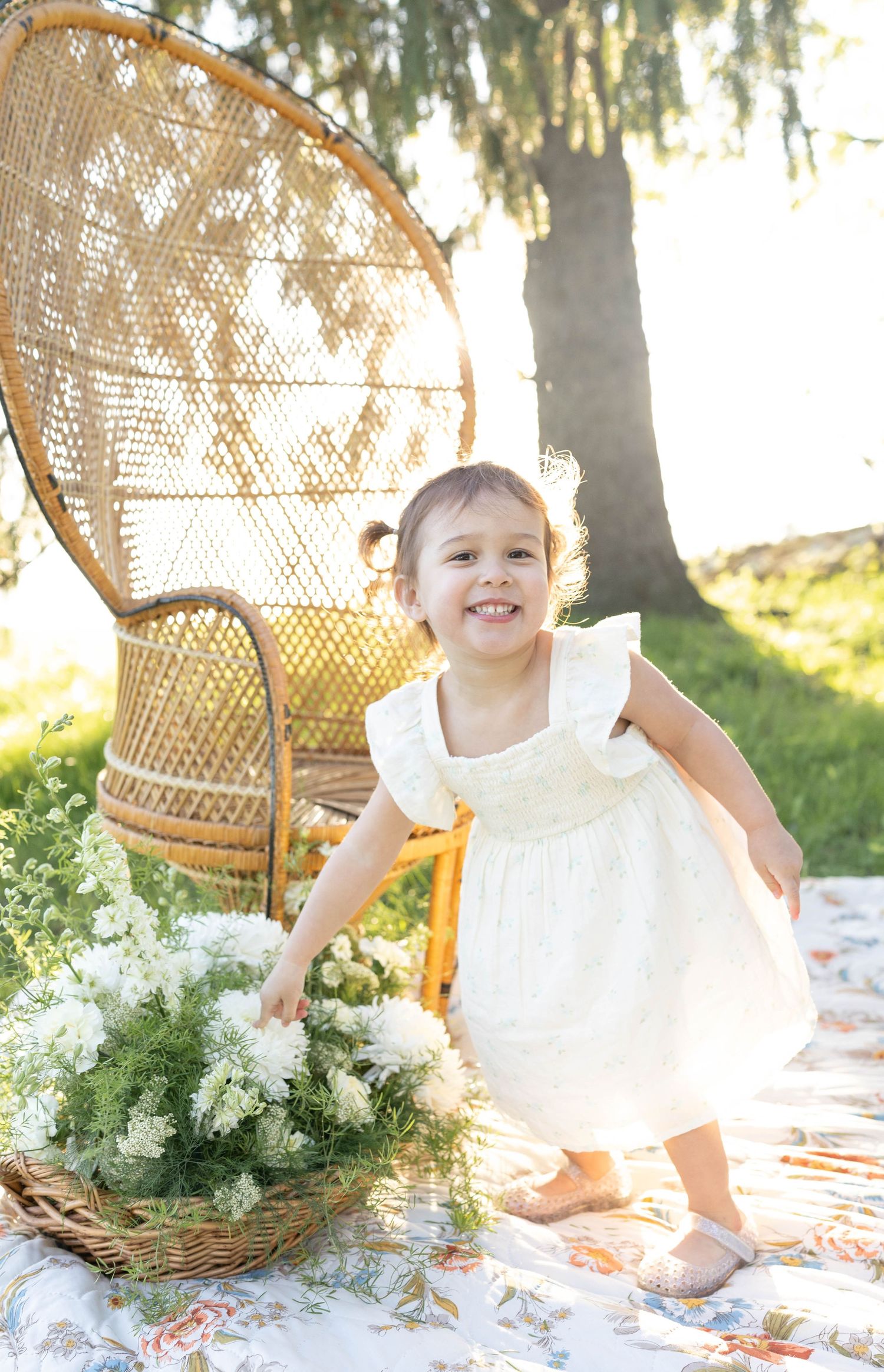 A child in a white dress stands in a sunny garden near a wicker peacock chair and floral basket.