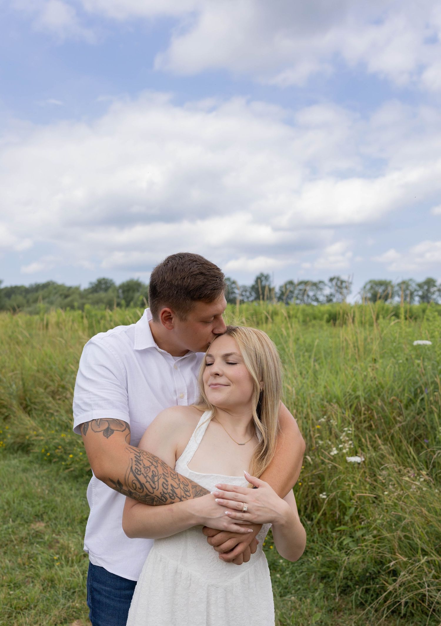 Tattooed man kissing woman's head while embracing her in a green meadow under a cloudy sky.