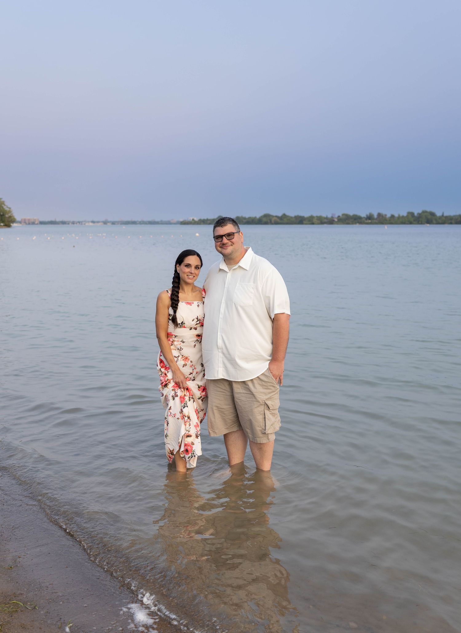 Couple standing in shallow lake water, woman in floral dress and man in white polo shirt and khaki shorts, smiling.