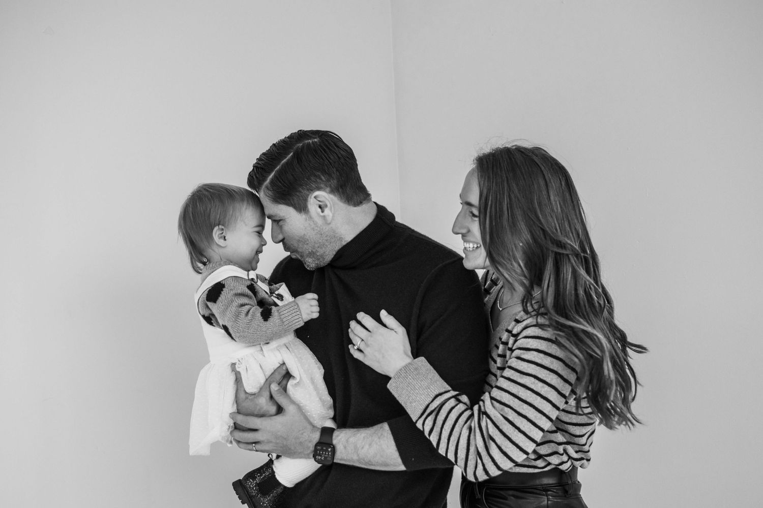 A black and white family portrait of parents sharing a playful moment with their baby against a plain wall.