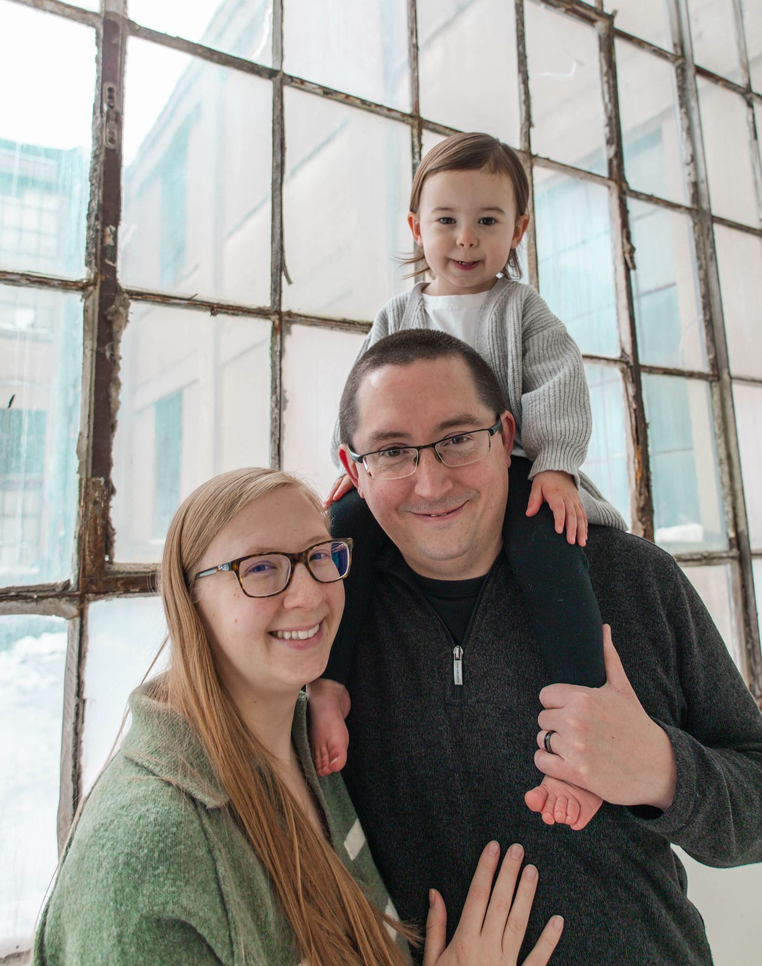 Happy family of three posing together by a large window in casual attire with natural lighting.
