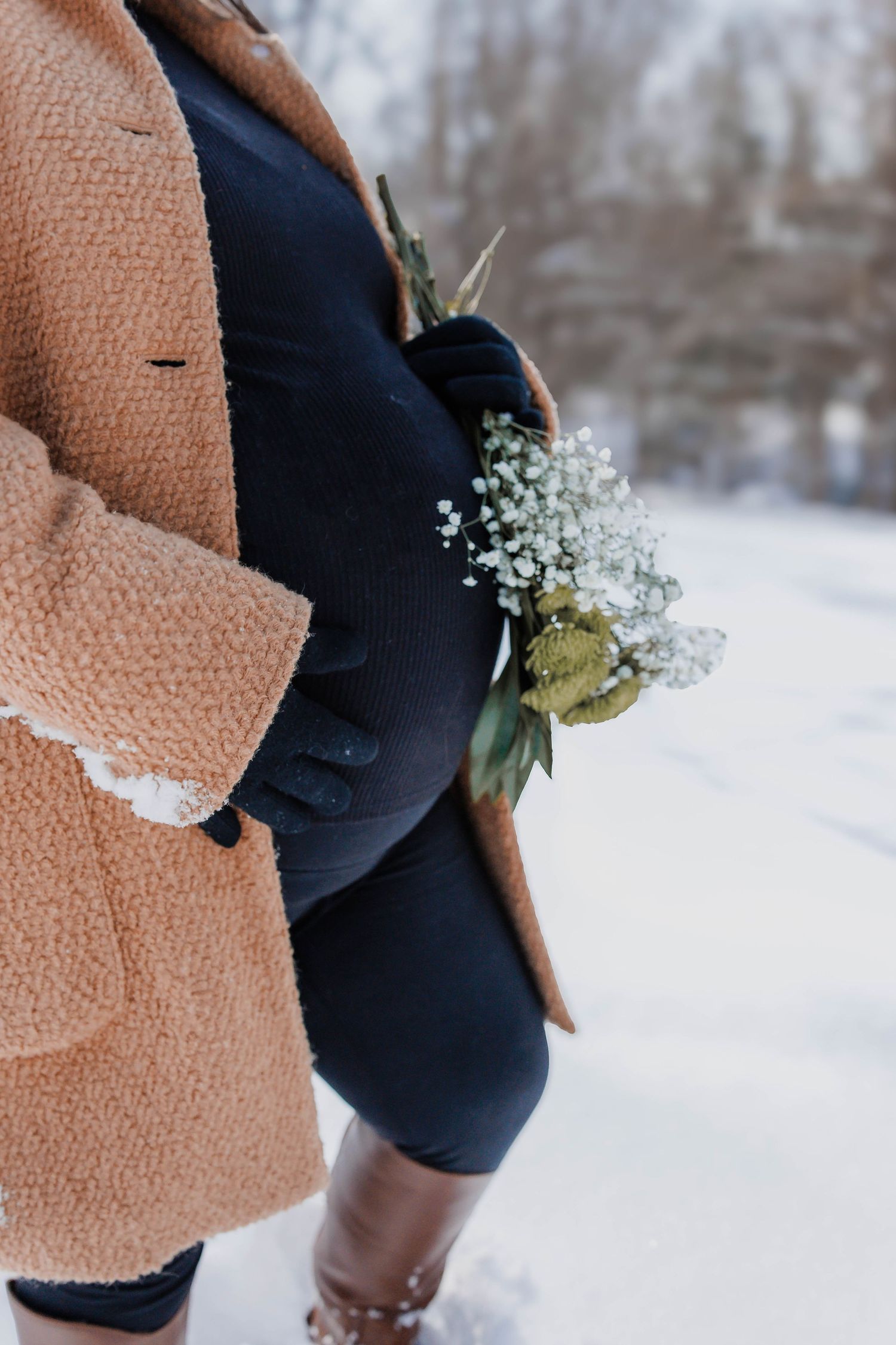 Pregnant woman in winter attire holds delicate baby's breath flowers against her belly in snowy setting.