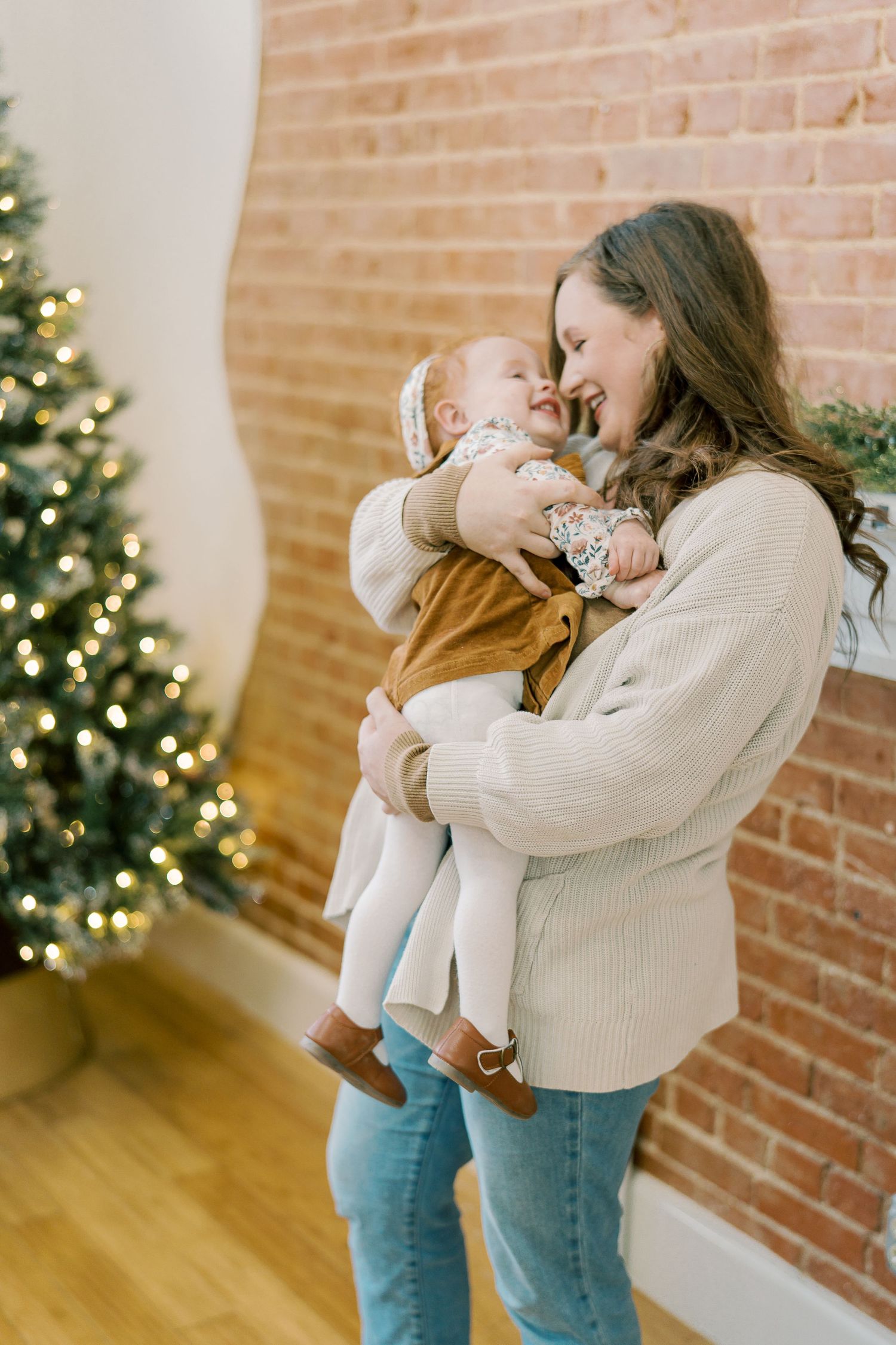 A tender moment shared between parent and baby in front of a decorated Christmas tree and exposed brick wall.