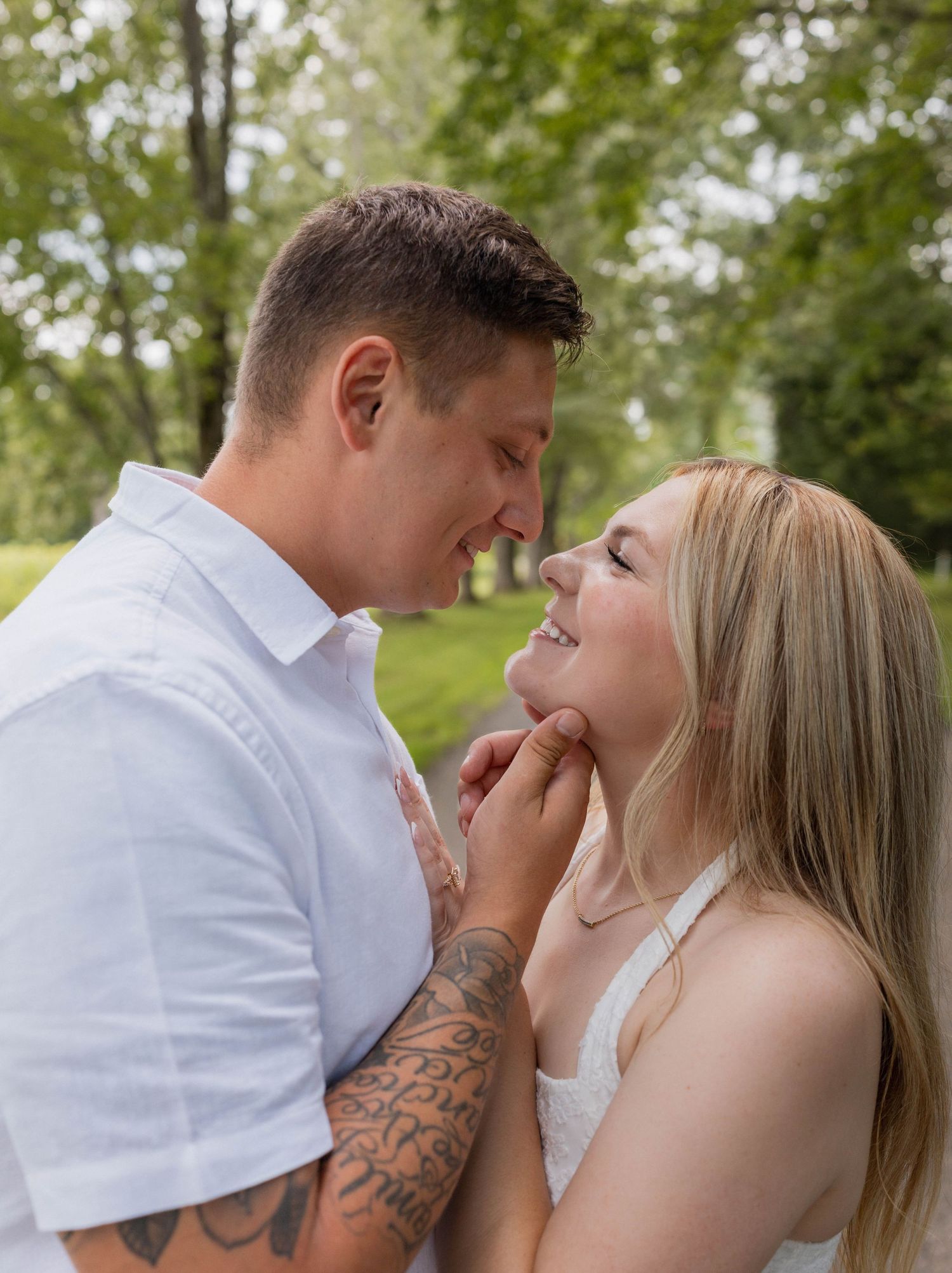 A couple shares a romantic moment outdoors, the man gently holding the woman's chin, both smiling, surrounded by lush green trees.