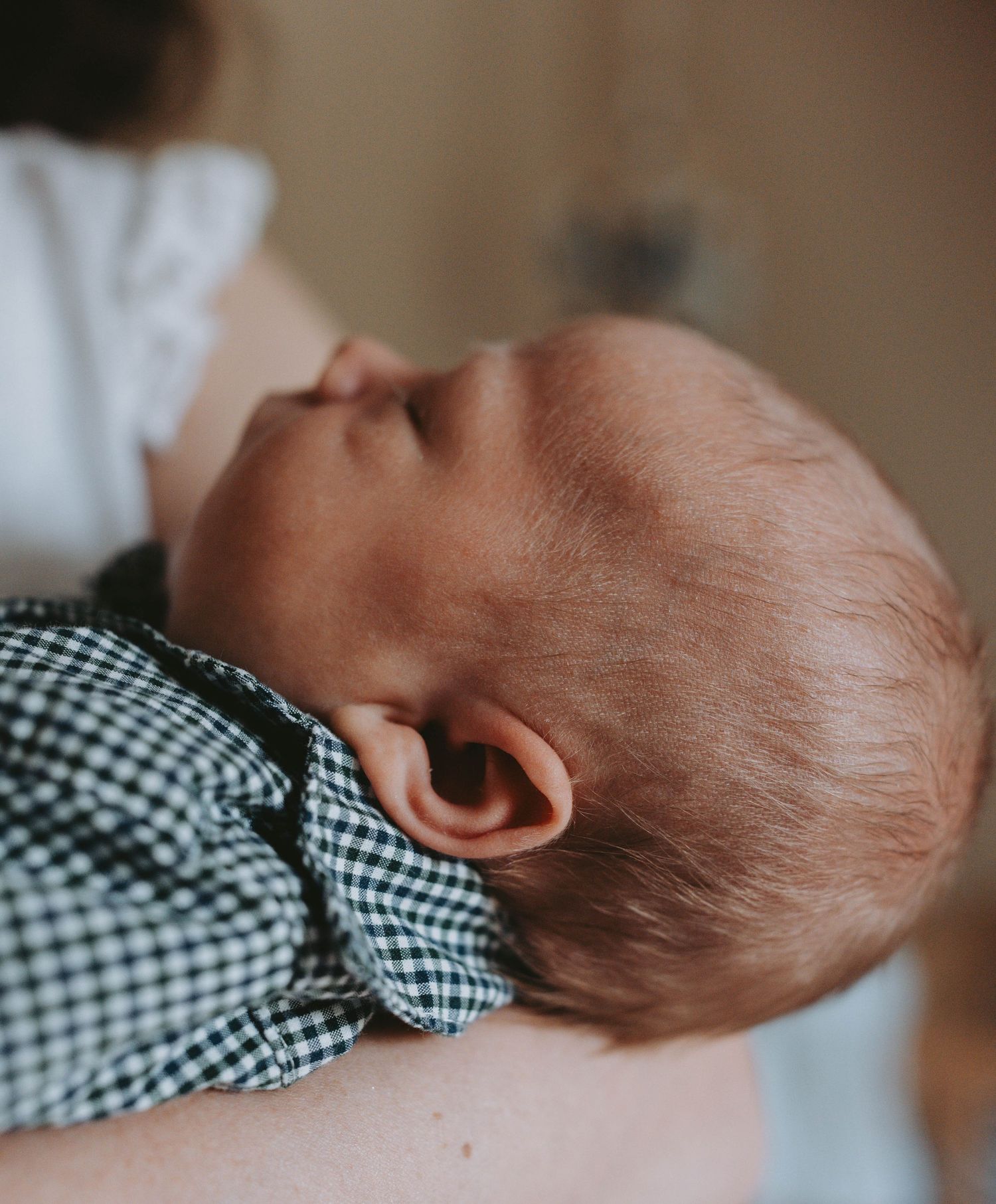 Newborn baby with red hair sleeping peacefully in a parent's arms wearing a gingham outfit.