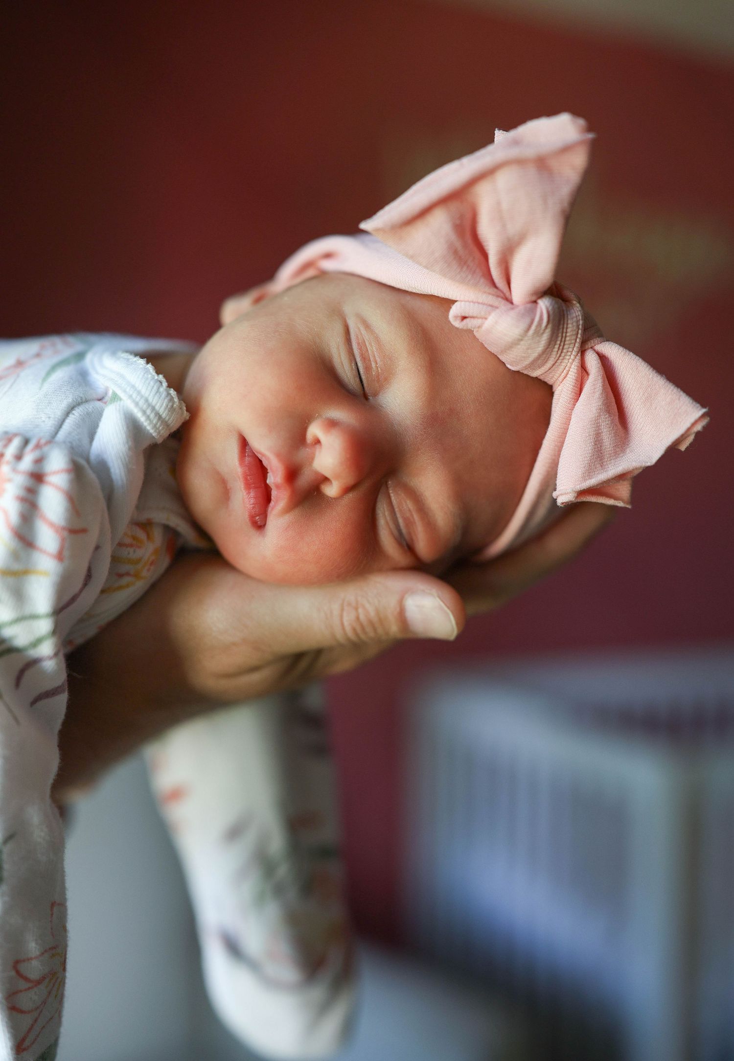 Sleeping newborn baby girl wearing a pink bow headband, cradled gently in an adult's hands.