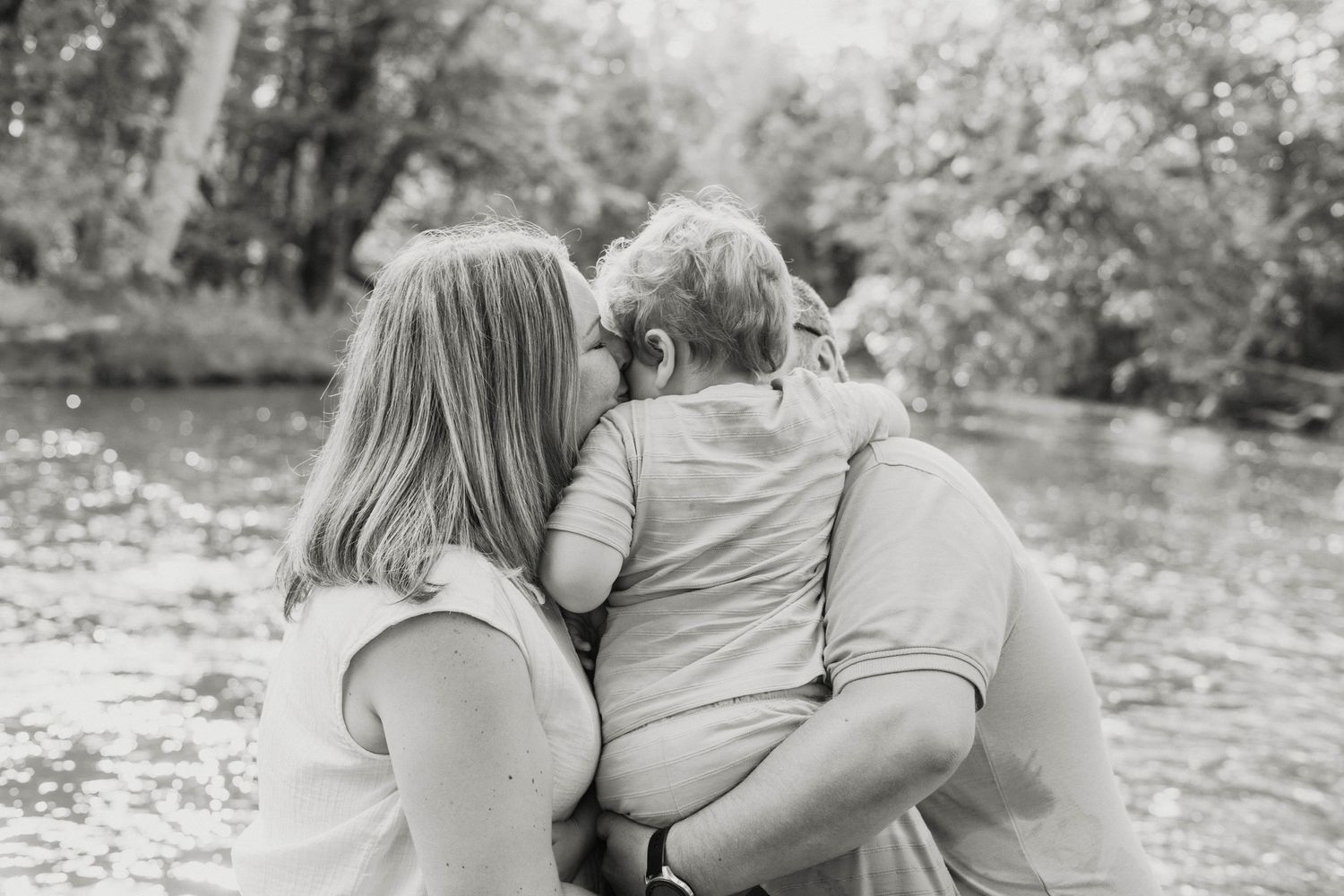 Black and white photo of a family embracing near a peaceful pond surrounded by lush green trees.