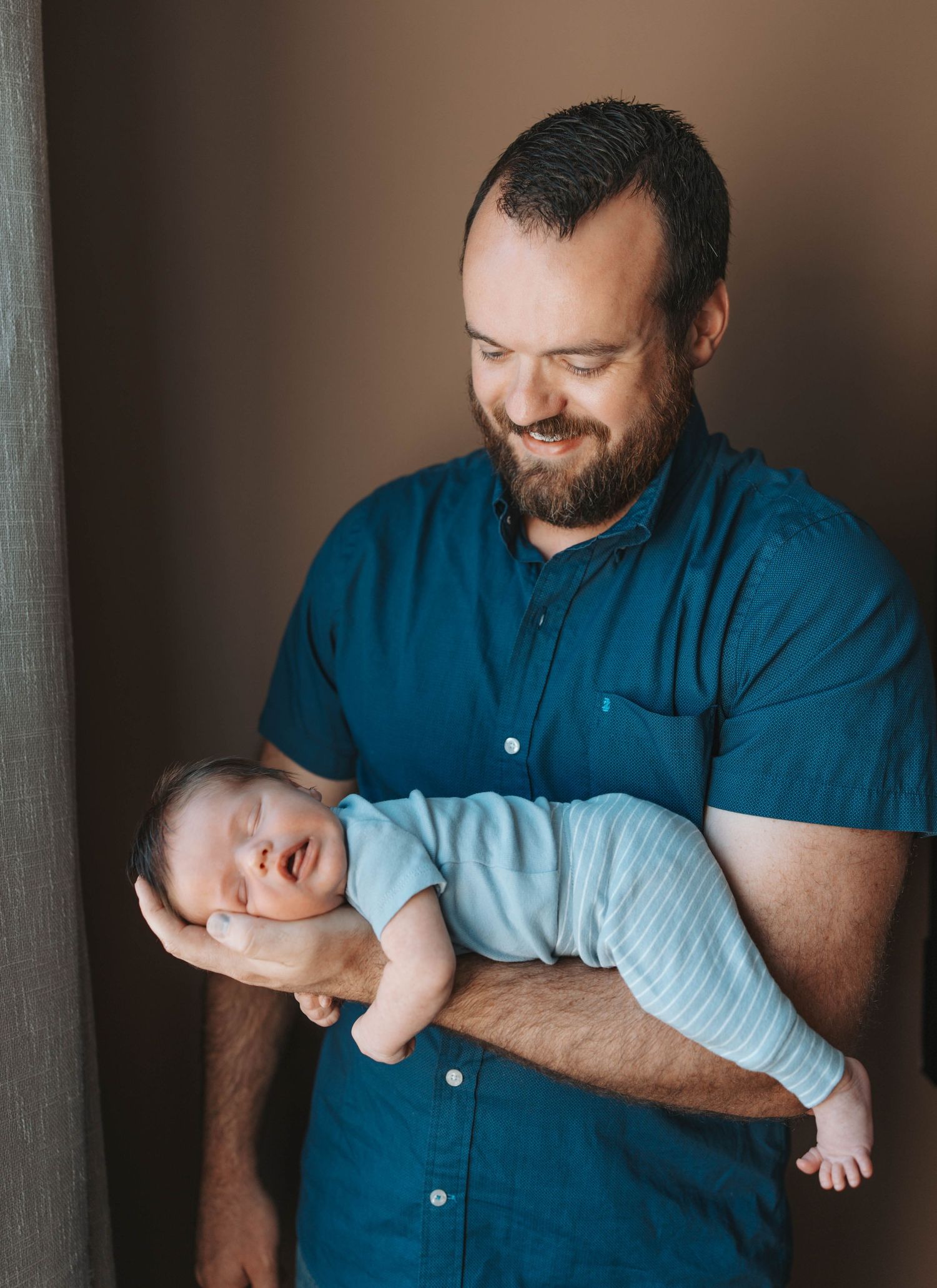 Smiling bearded man in teal shirt cradling a sleeping newborn baby in his arms indoors.