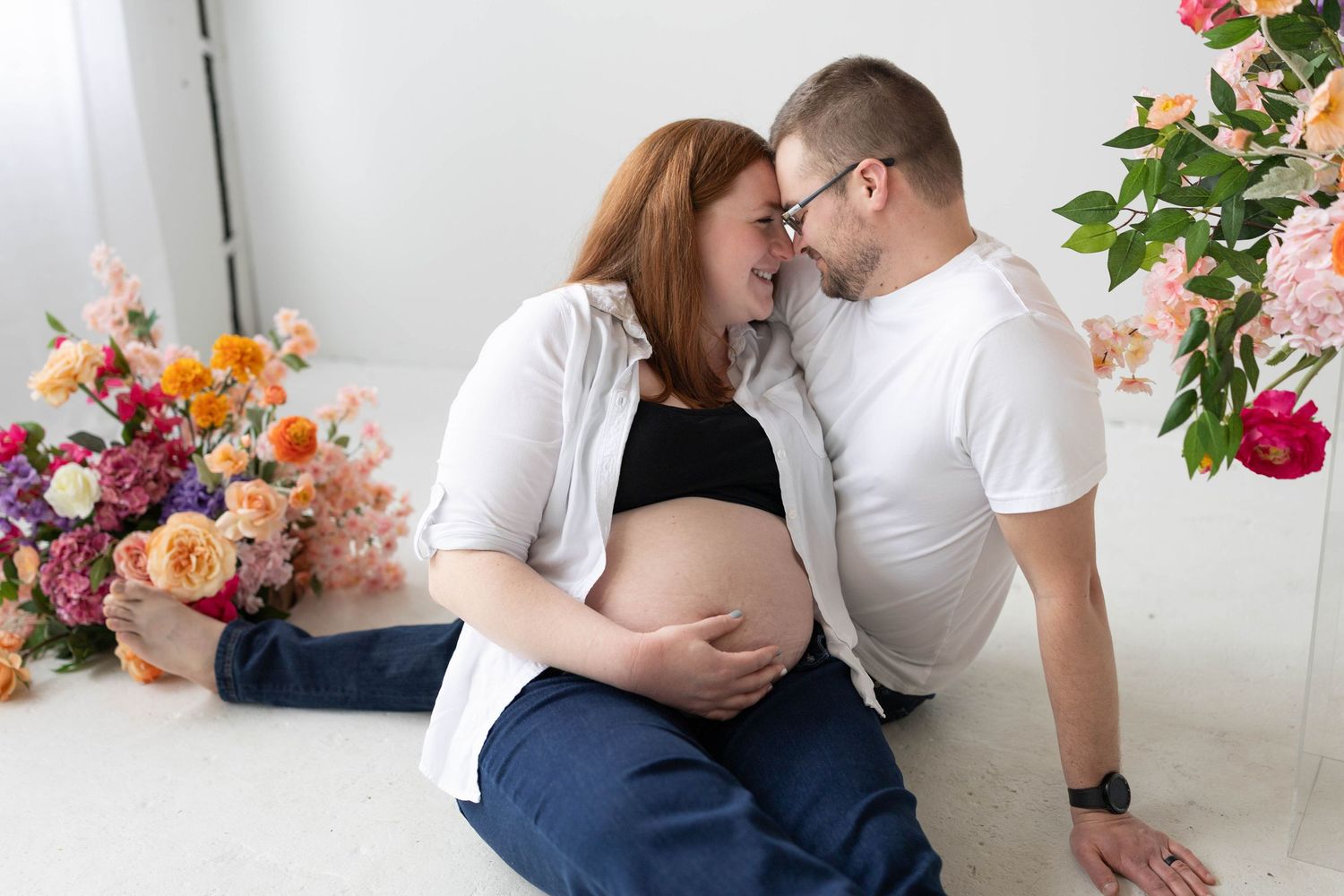 Expecting couple shares intimate moment surrounded by colorful floral arrangements in bright studio setting.