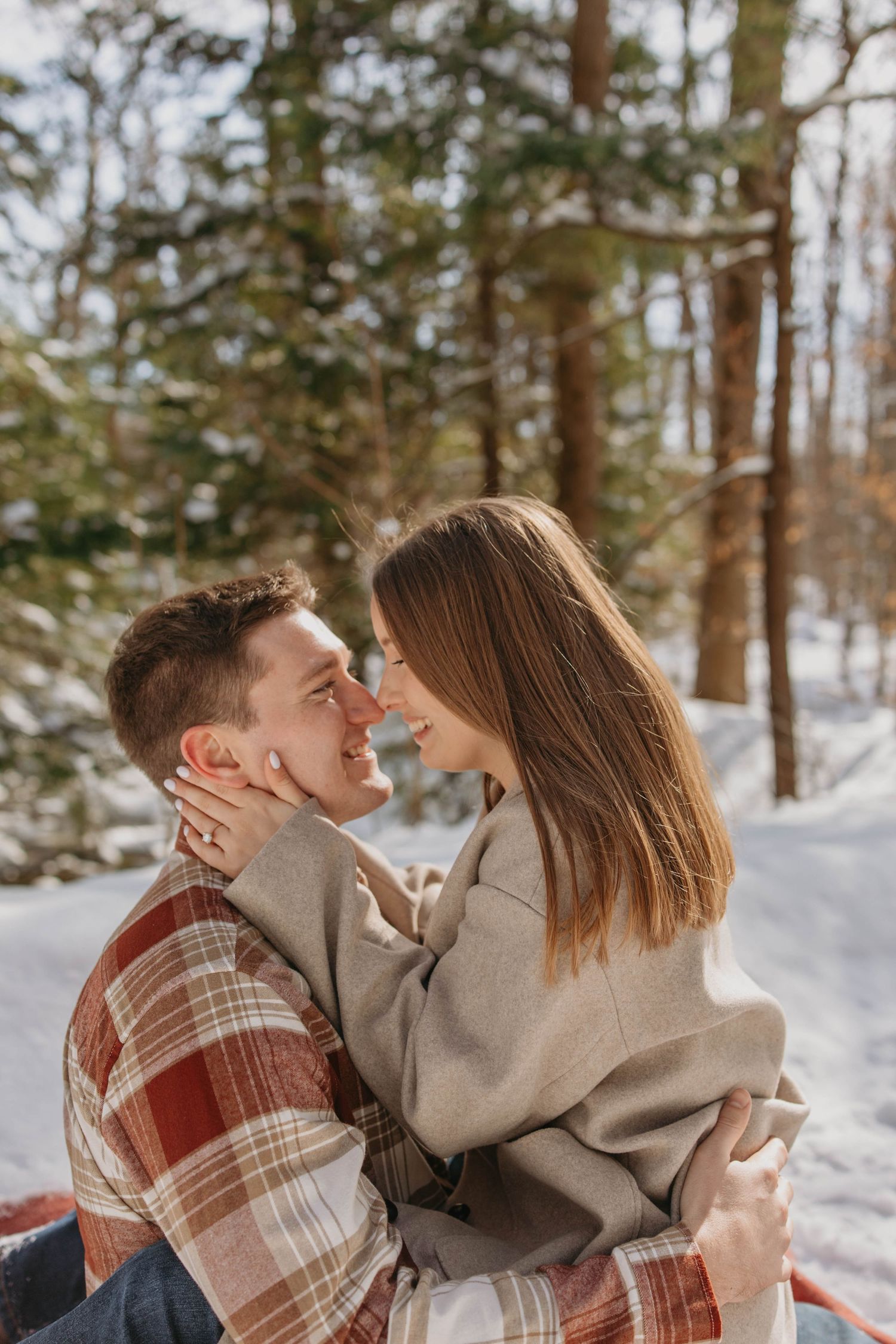 A couple in cozy winter clothing share a romantic moment together in a snowy forest setting.