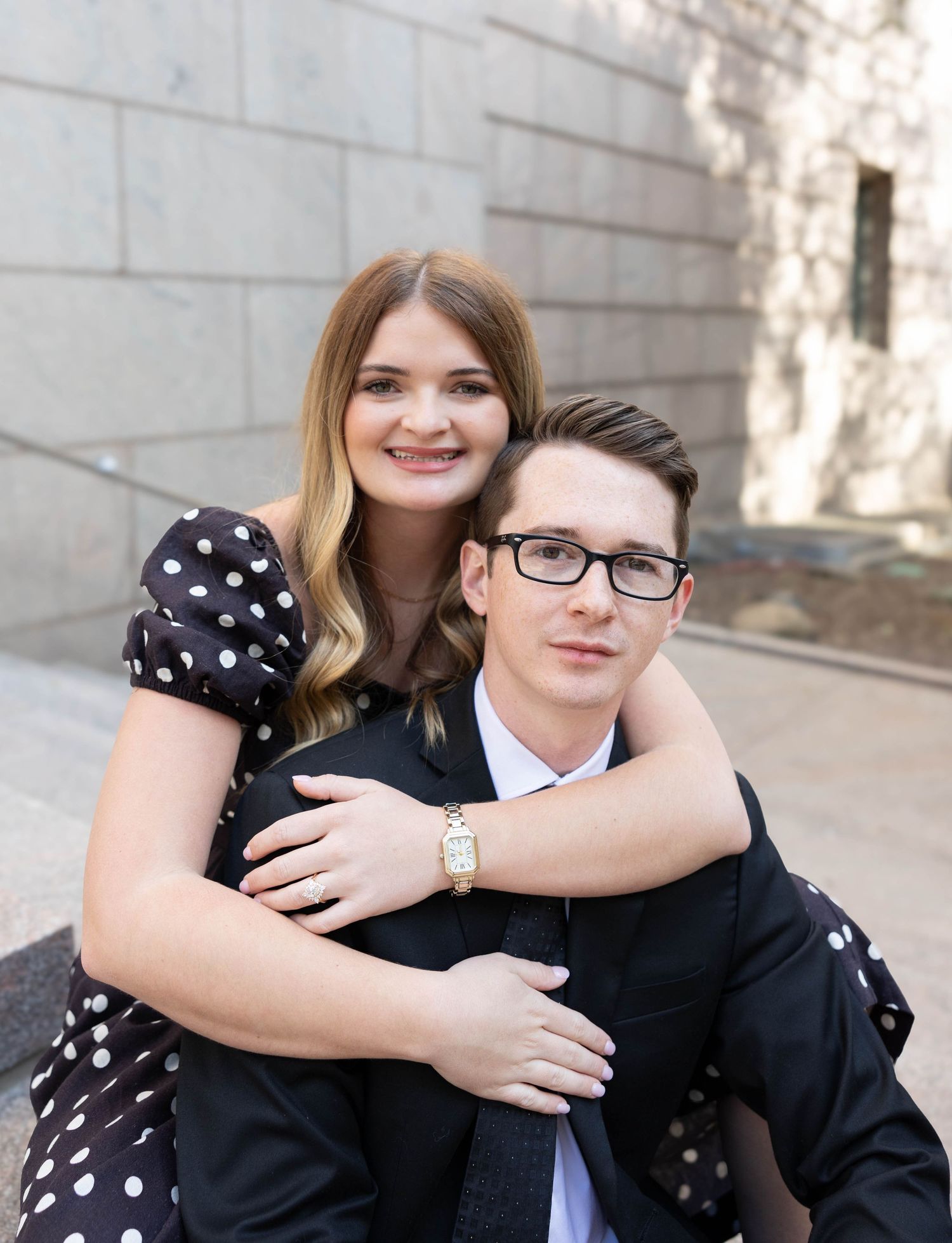 A couple poses together outdoors, with one wearing a polka dot dress and the other in a dark suit with glasses.