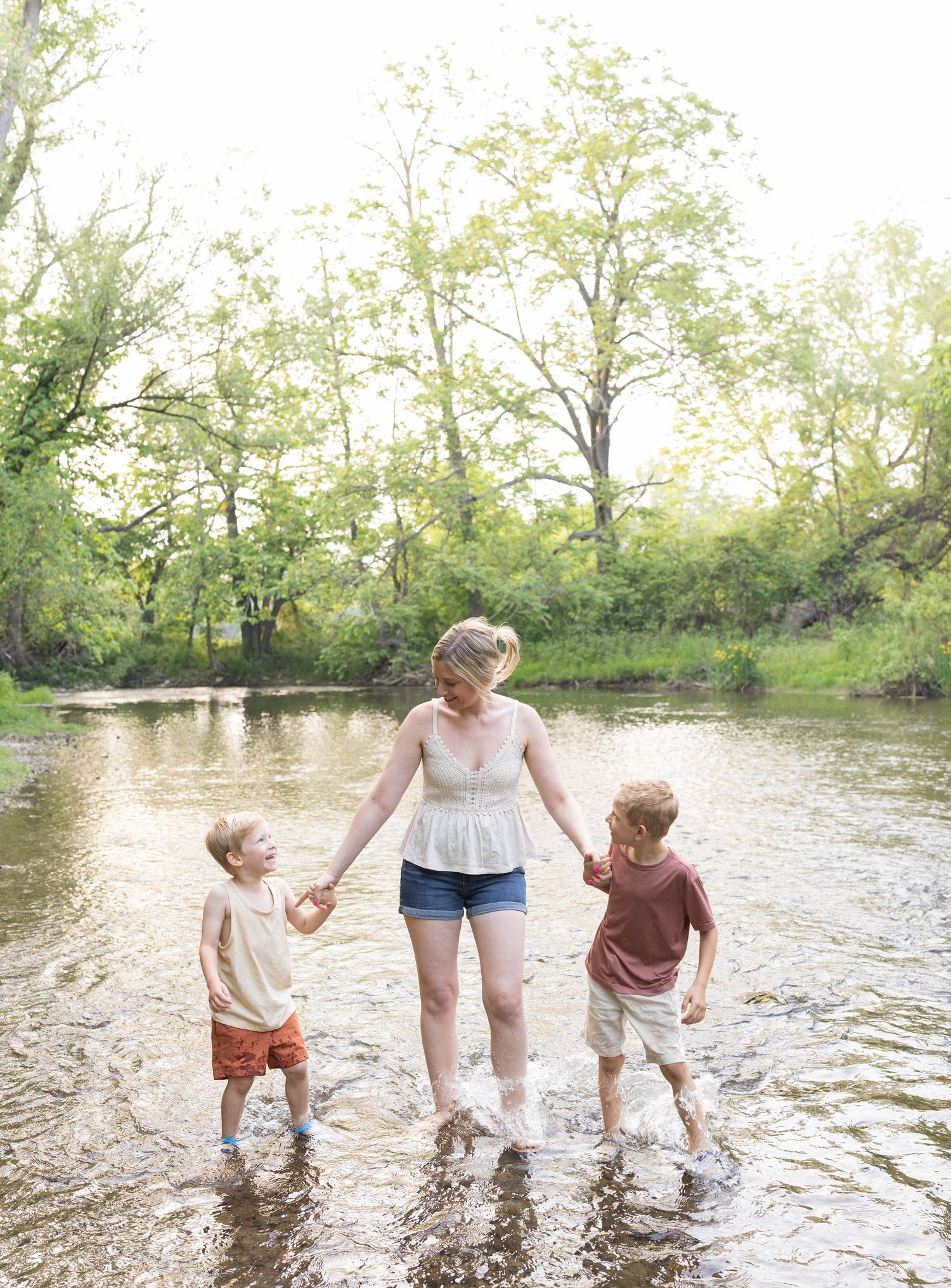 Three people splash together in a shallow creek on a warm summer day with lush green trees in the background.