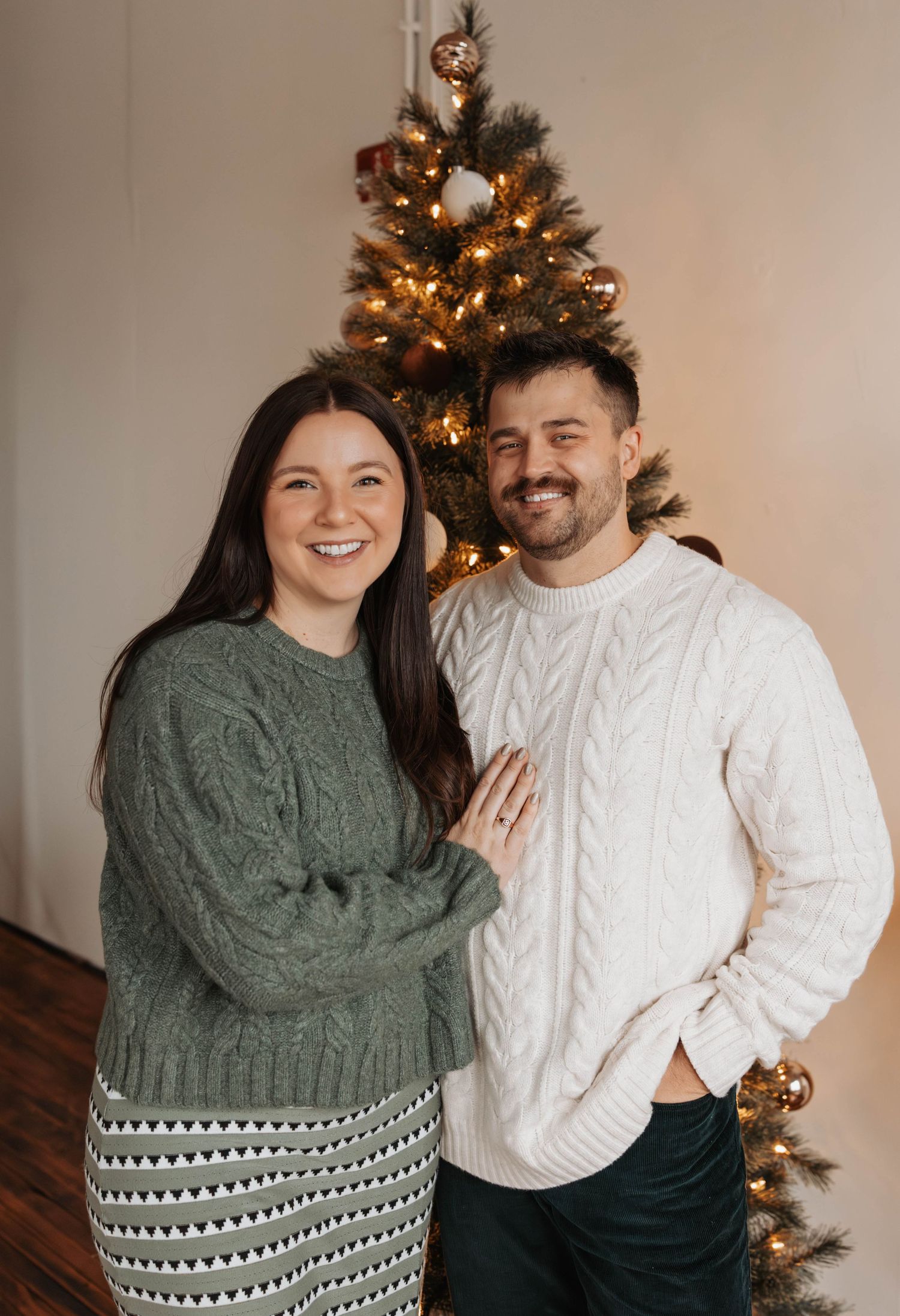 A couple poses together in front of a Christmas tree in a series of candid holiday photos wearing cozy winter sweaters.