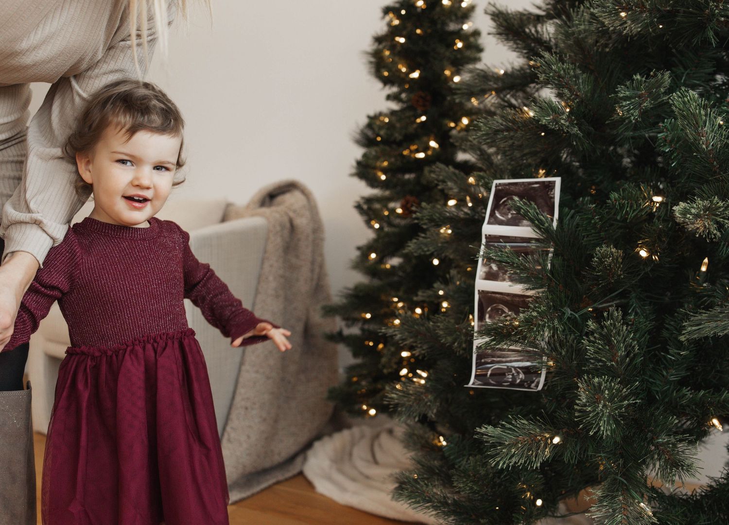 A young child in a burgundy dress twirls and plays in front of a decorated Christmas tree and holiday decor.