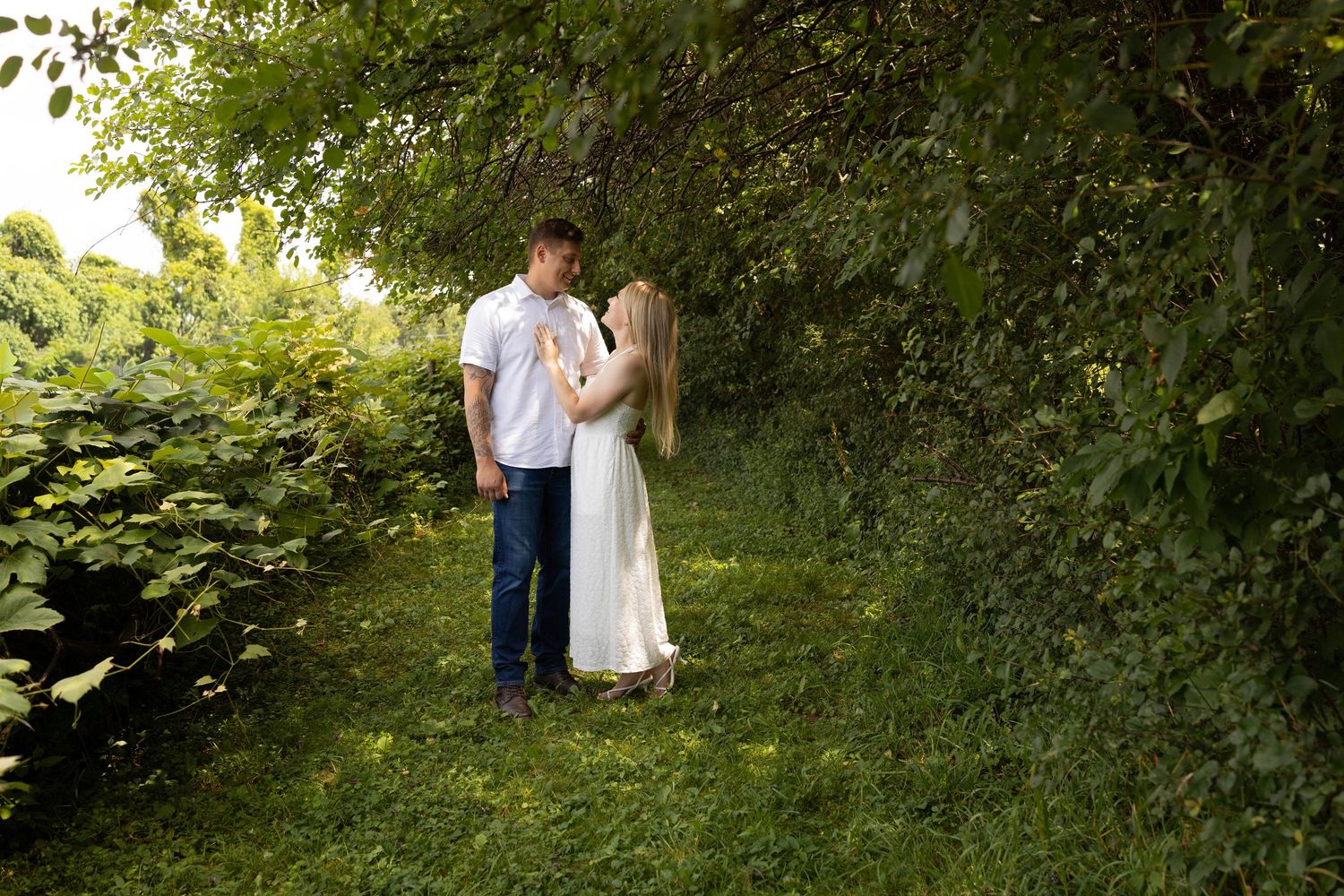 Couple sharing a romantic moment in a lush green garden pathway, woman in white dress, man in jeans and white shirt.