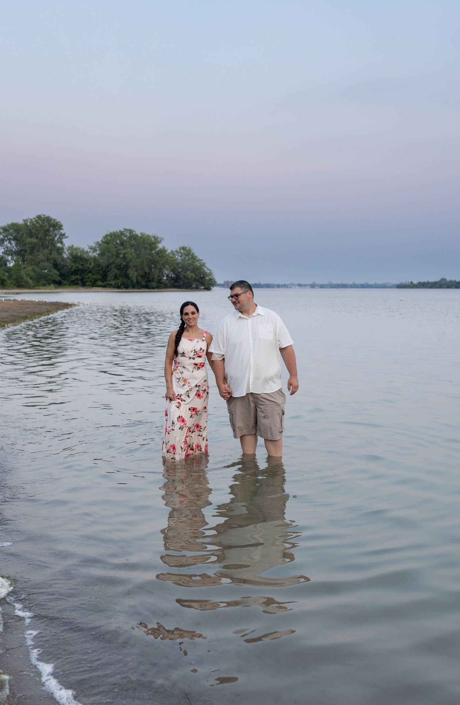 Couple holding hands wading in shallow water at sunset, woman in floral maxi dress, man in white shirt and shorts.