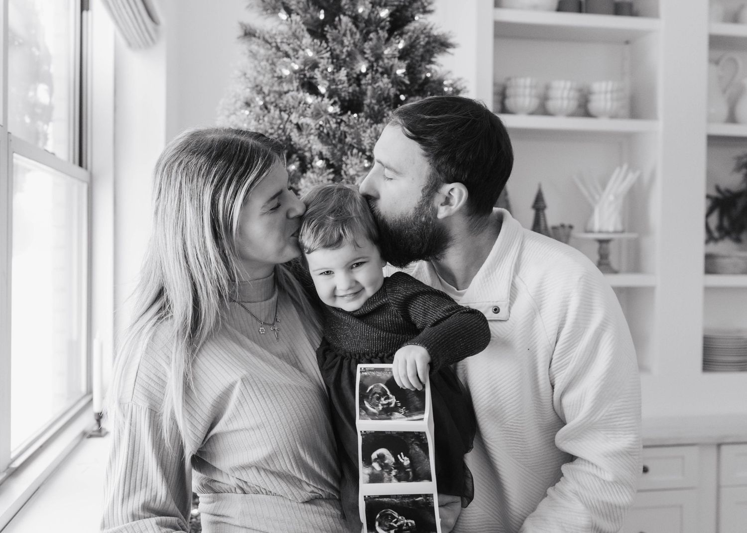 Black and white photo of parents kissing their toddler while holding ultrasound photos in a bright kitchen setting.
