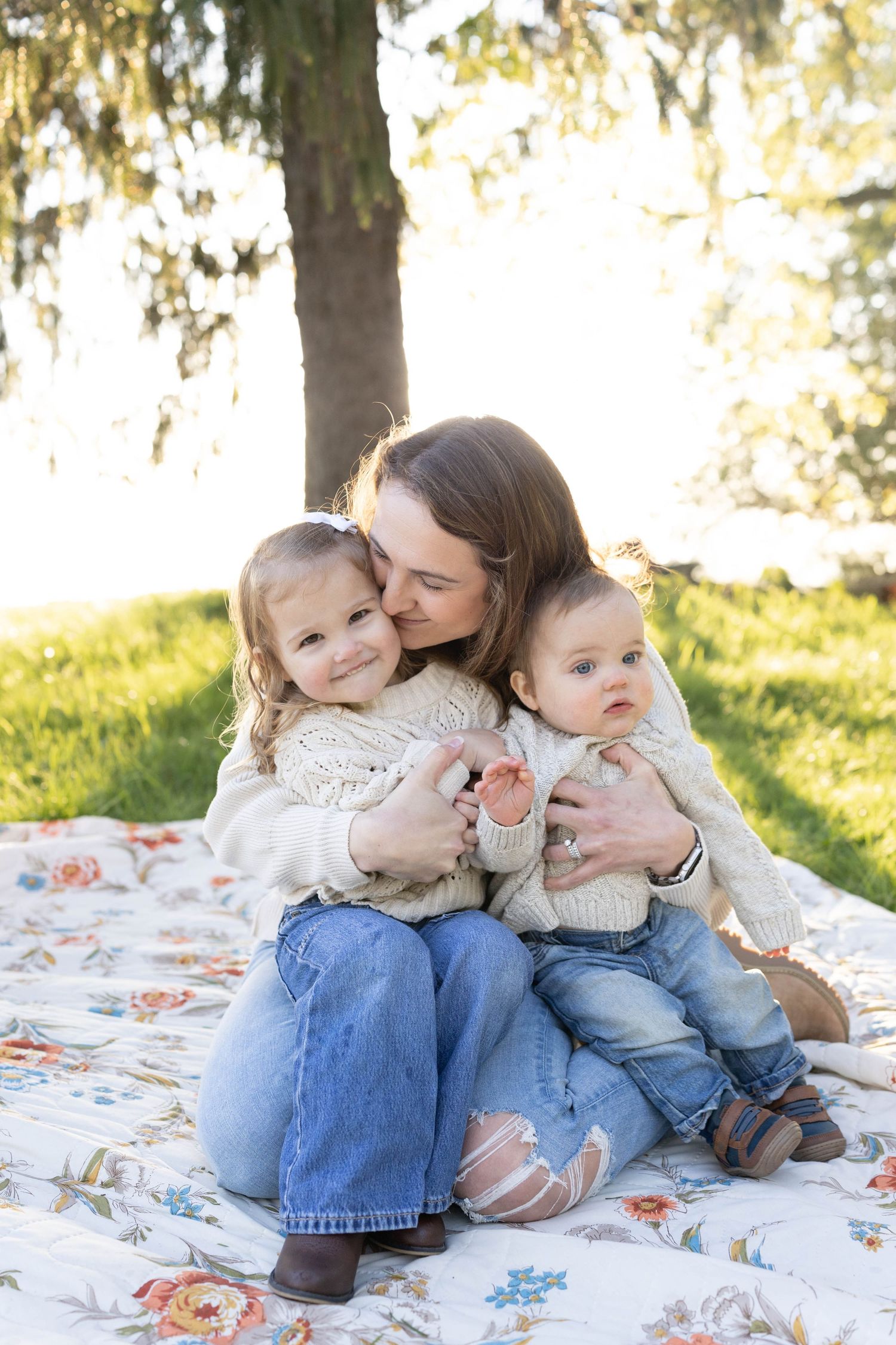 Family sits together on floral blanket during outdoor photo session in sunlit park setting.
