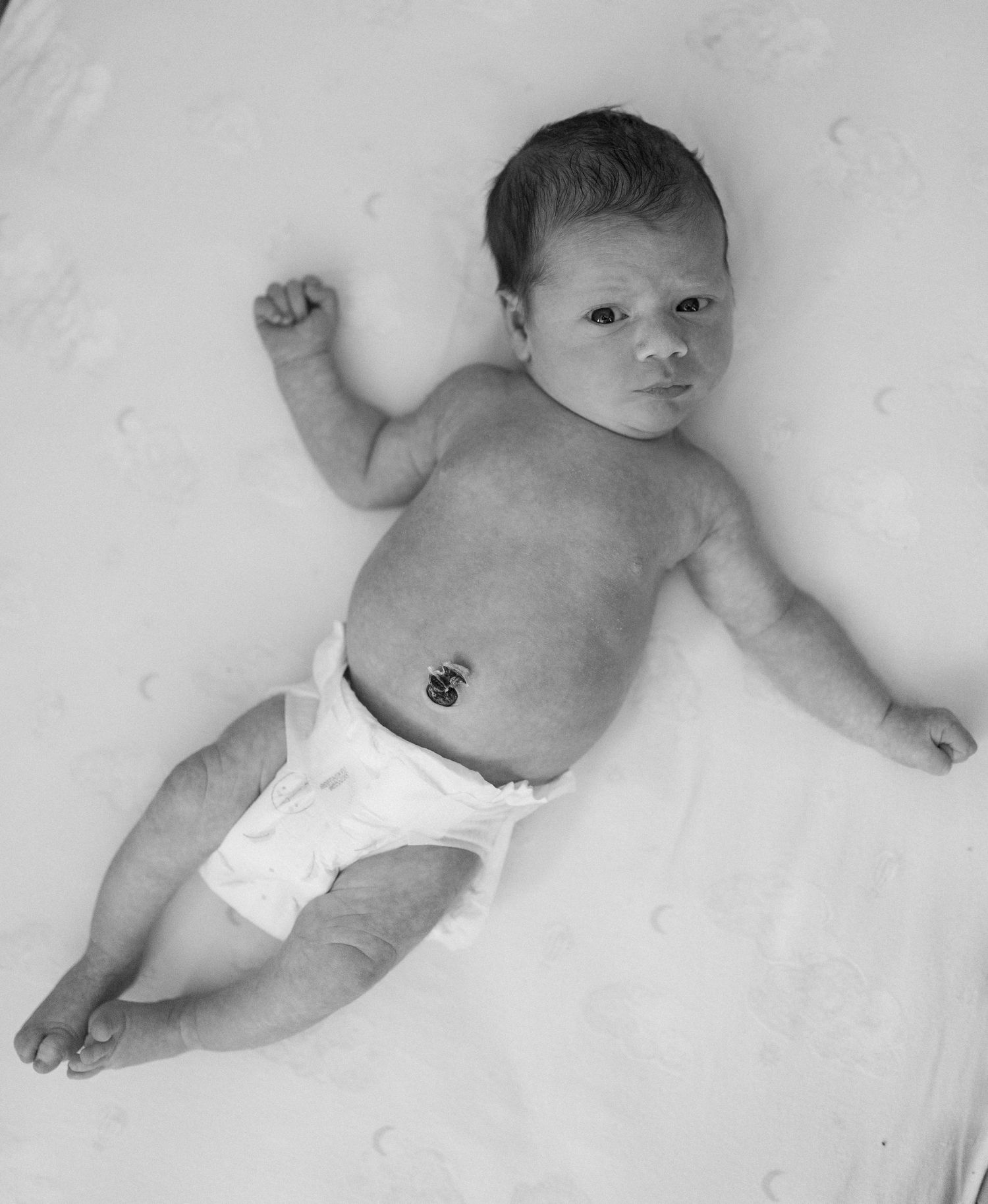 Black and white photo of a baby wearing a diaper lying on a light background with one arm raised.