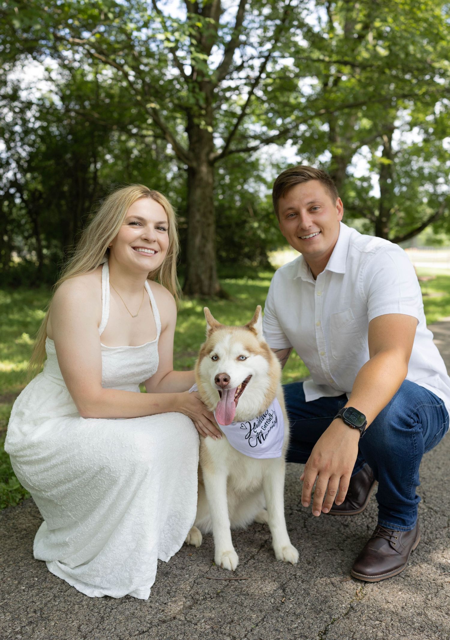 A smiling couple in white dress and shirt pose with a Husky dog wearing a bandana outdoors among green trees.