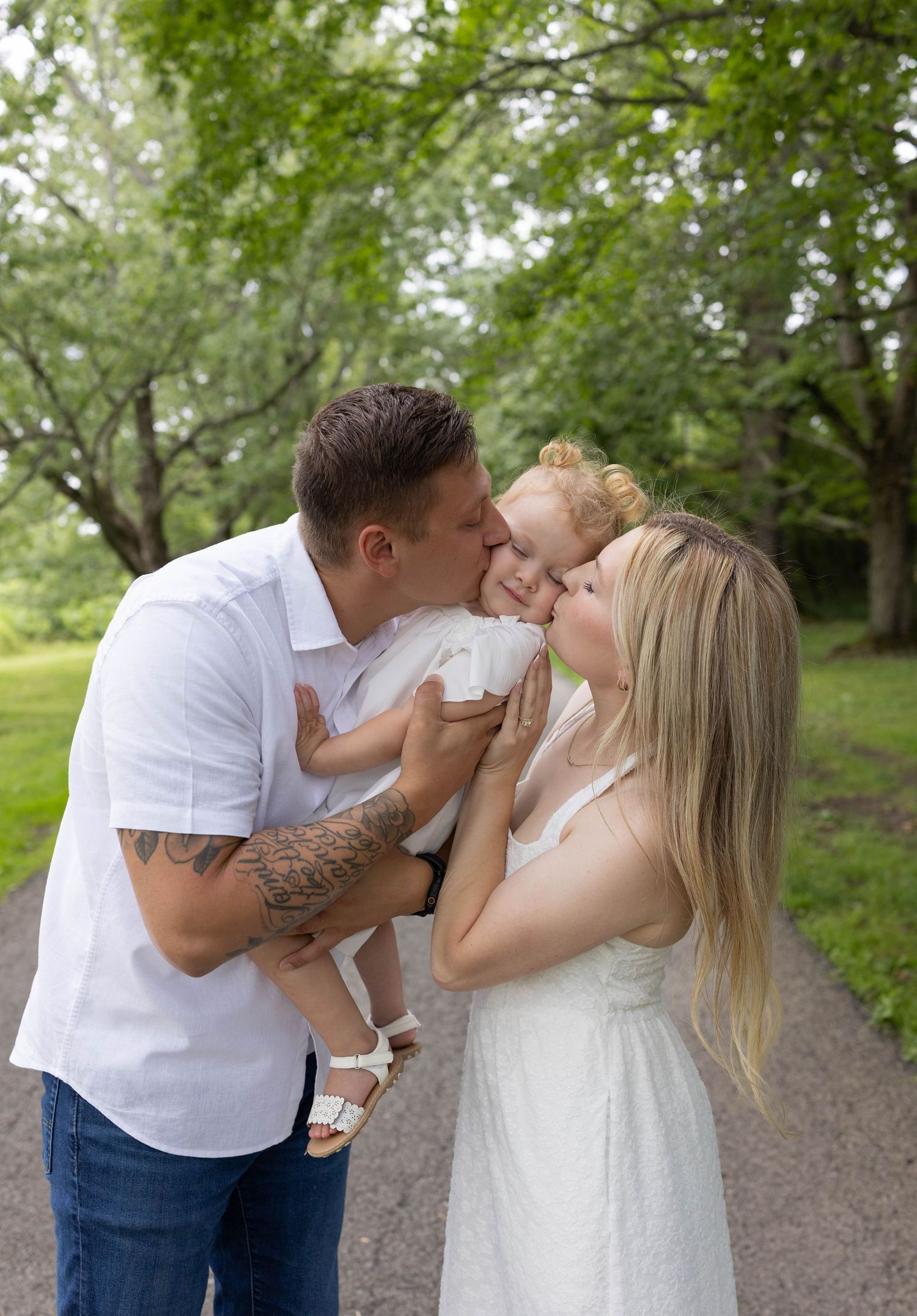 A young family shares a sweet moment as both parents kiss their toddler on a tree-lined path.