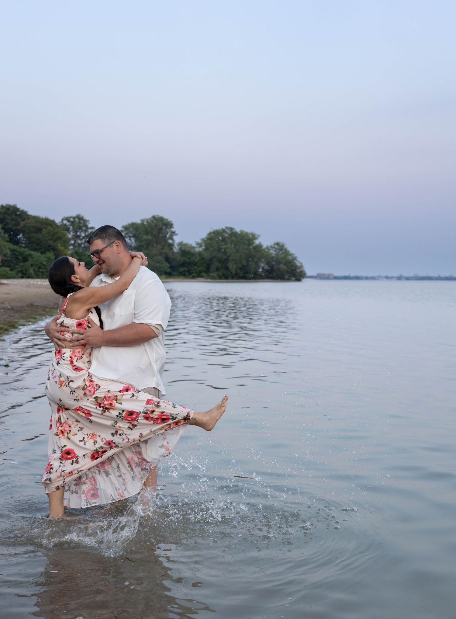 Couple embracing and kissing while standing in shallow lake water at dusk, woman in floral dress lifted by man.