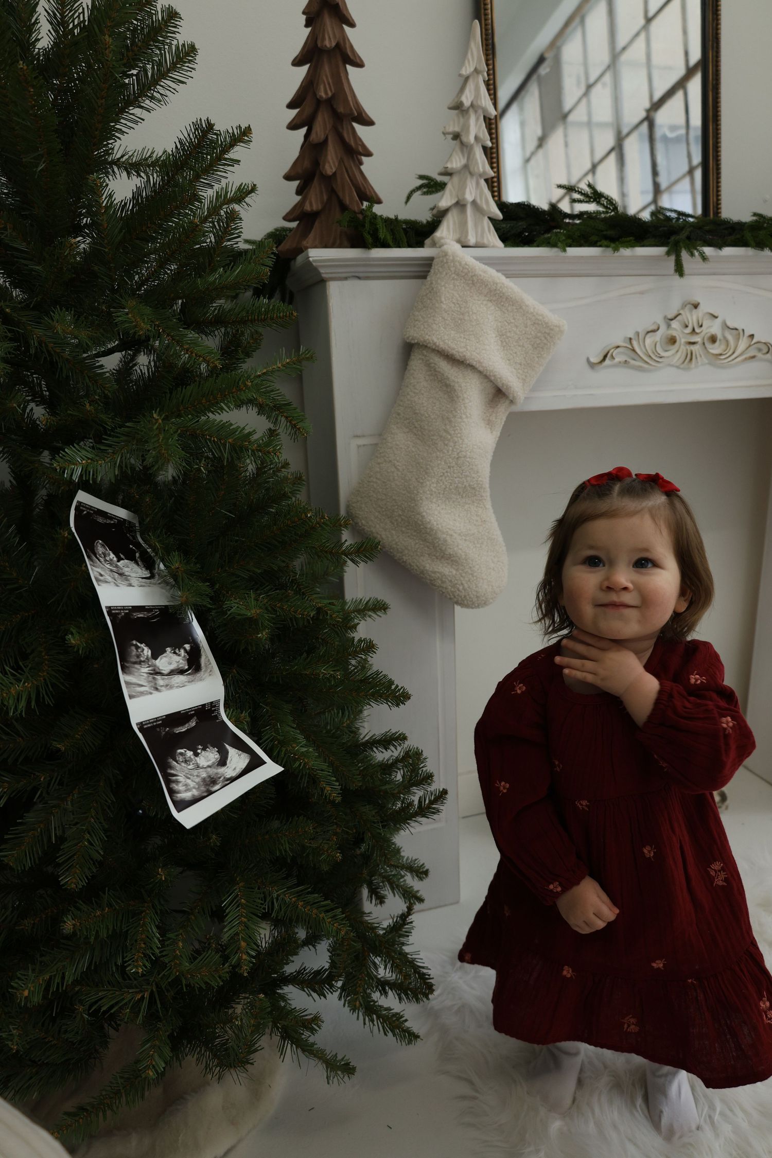 Holiday scene with Christmas tree, white stocking on mantle, and decorative wooden tree near a bright window.