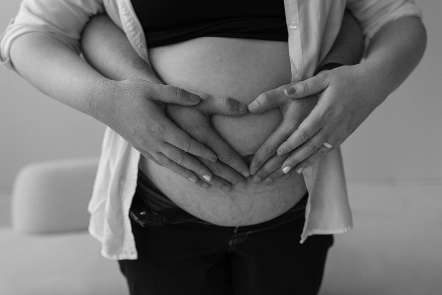 Black and white photo of hands forming a heart shape over a pregnant belly revealing bare skin under white cardigan.