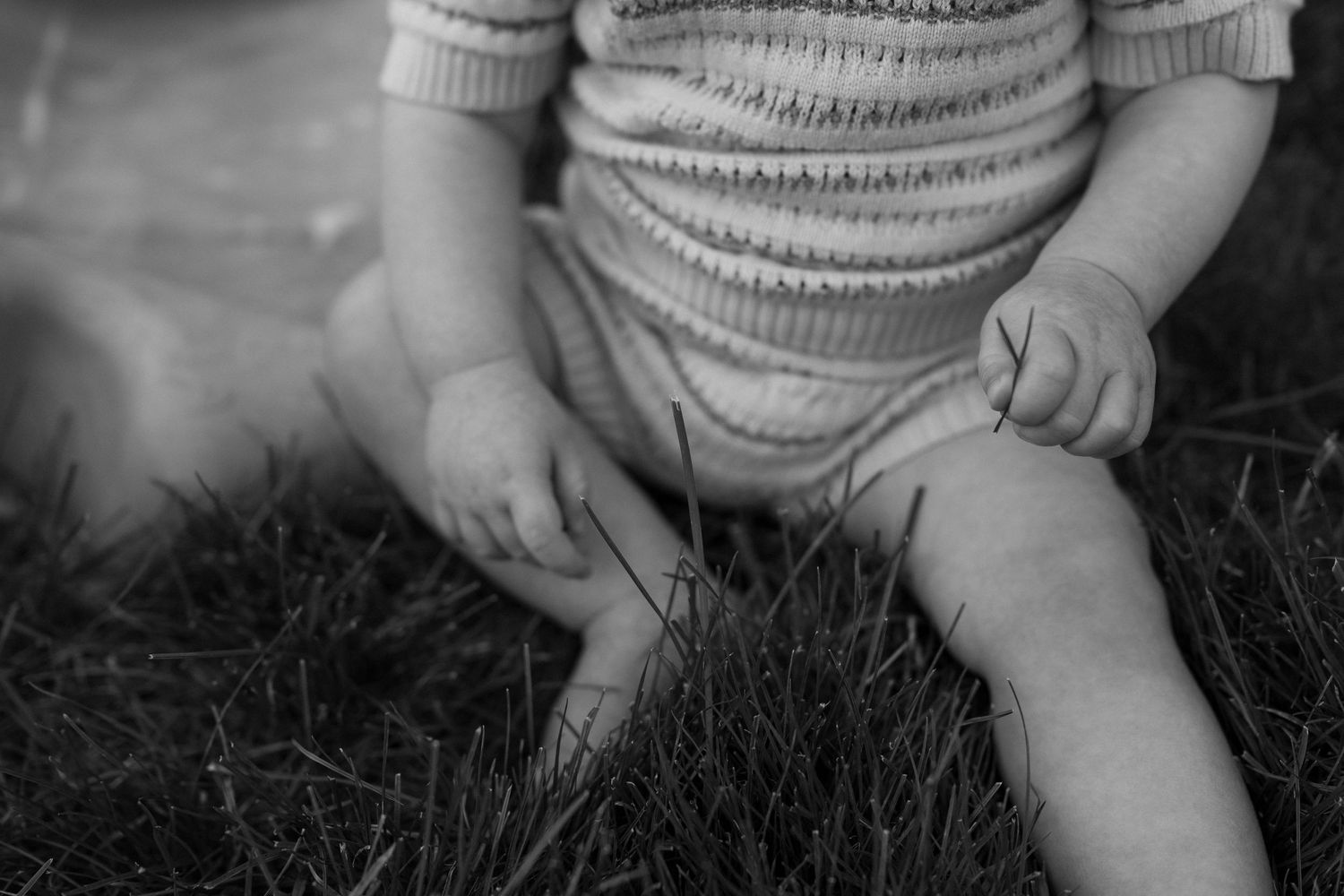 Baby wearing a knit sweater sits on grass while reaching down to touch something on the ground.