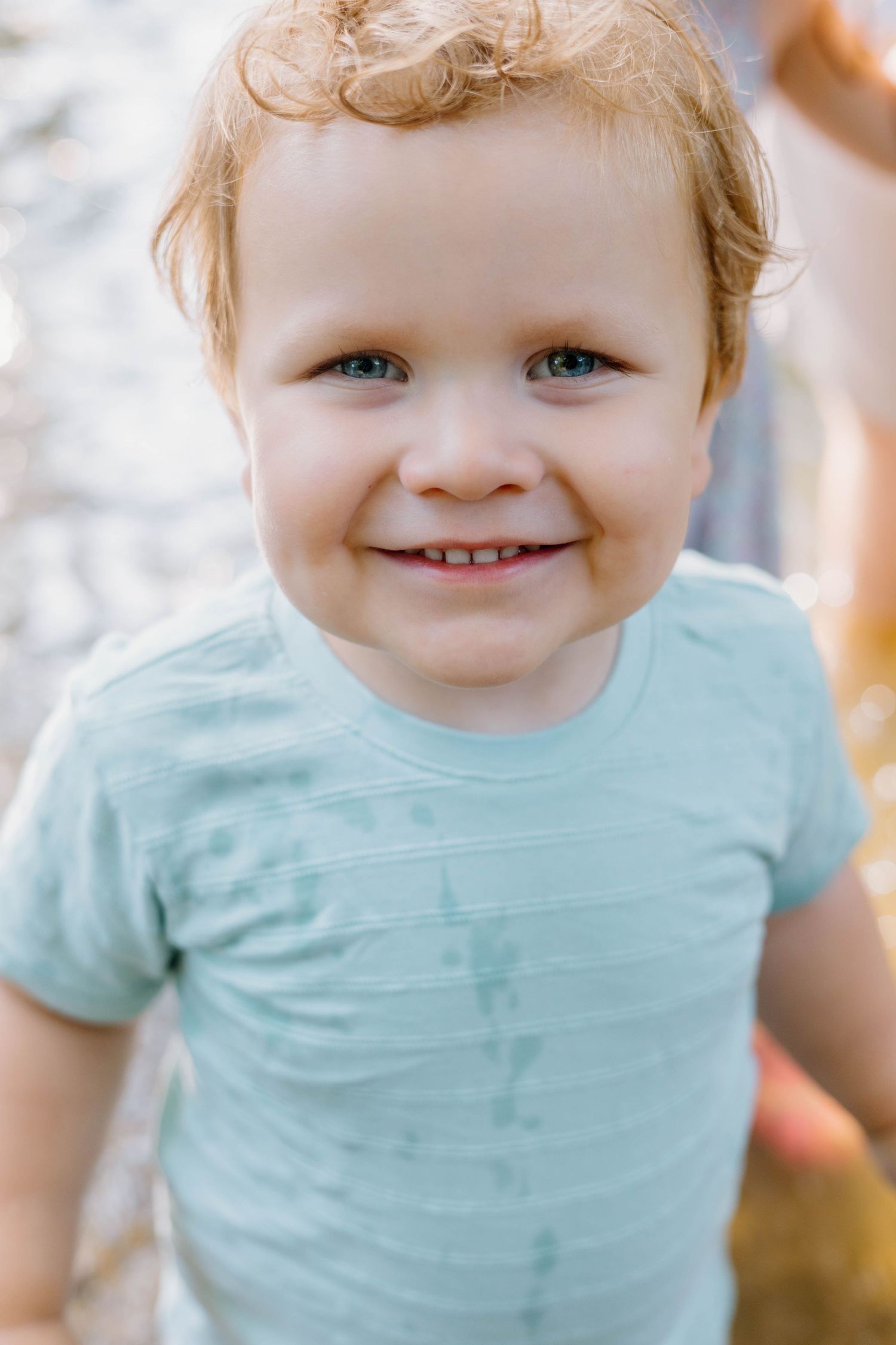 Smiling toddler with curly red hair and blue eyes wearing a light blue t-shirt outdoors.